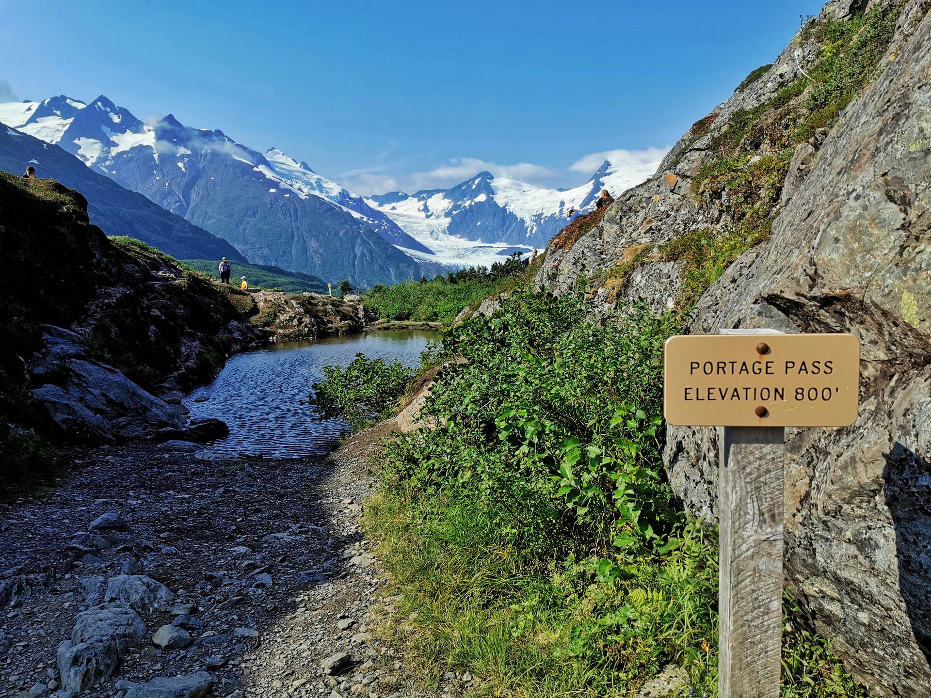 A trail sign stands beside a small pond, with snow-capped mountains in bright sunlight.