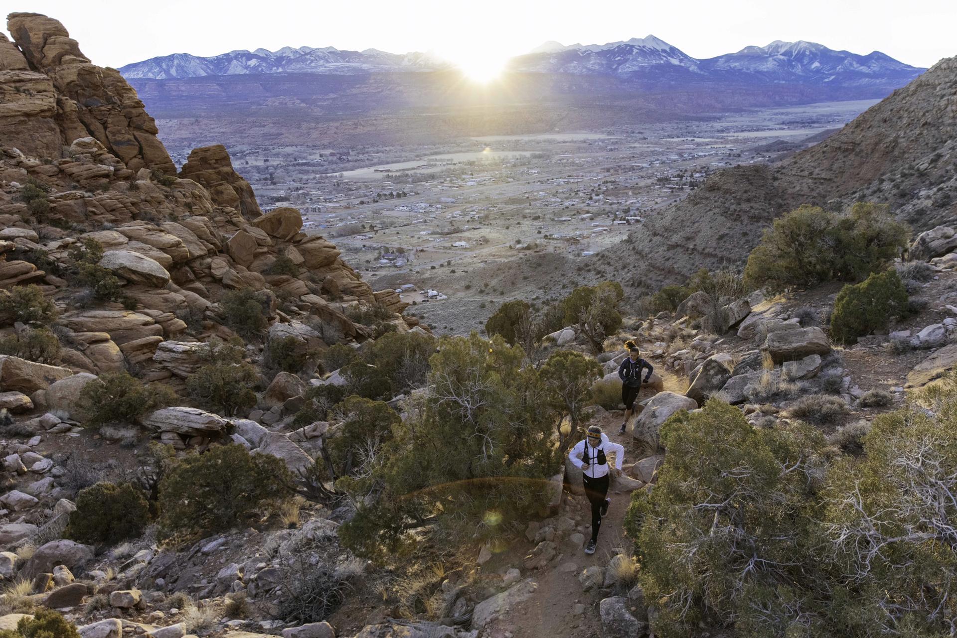 Two people run on a rocky trail at sunrise with mountains in distance.