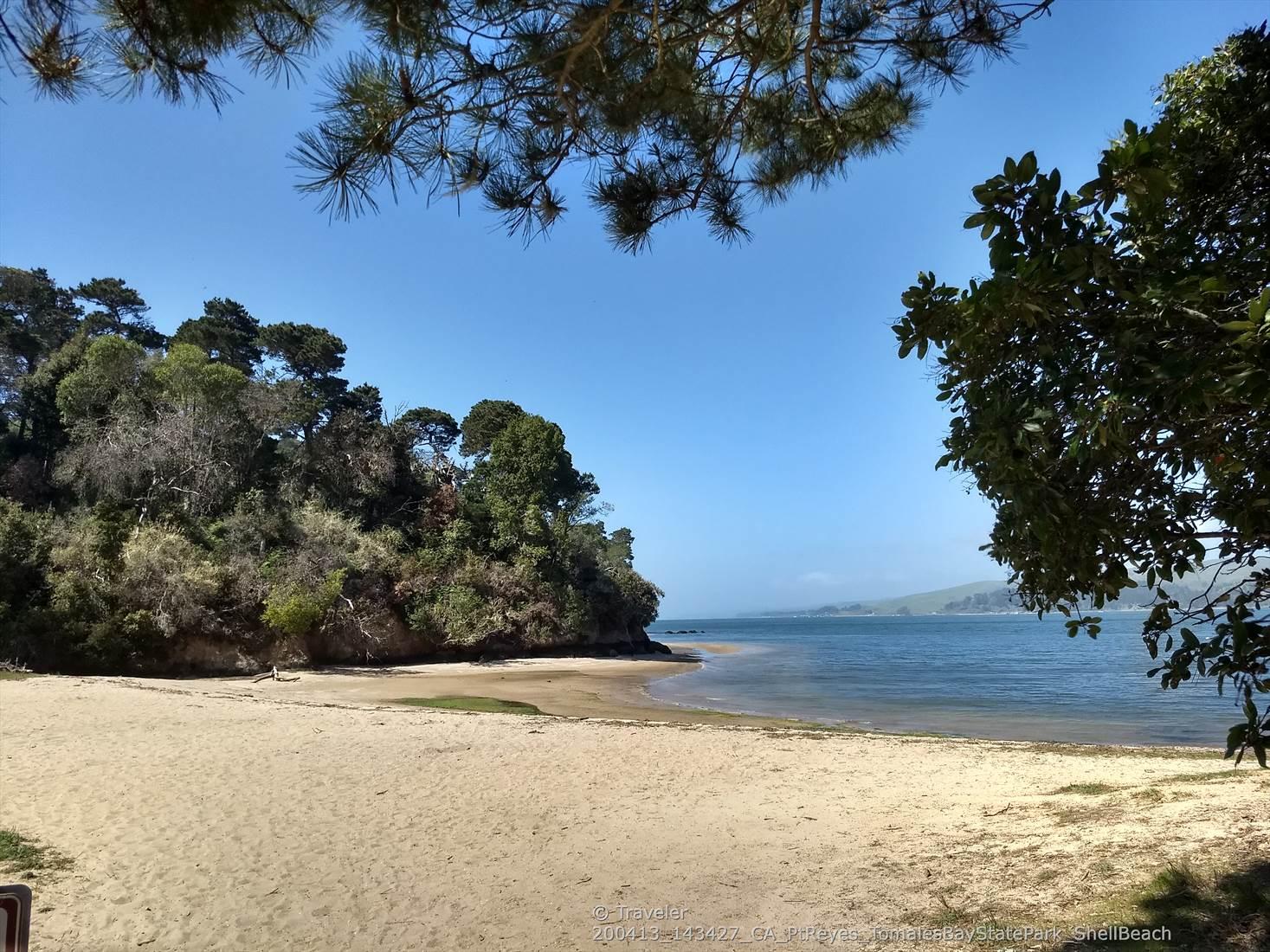 Wide sandy beach next to calm blue water and a green forested hill.