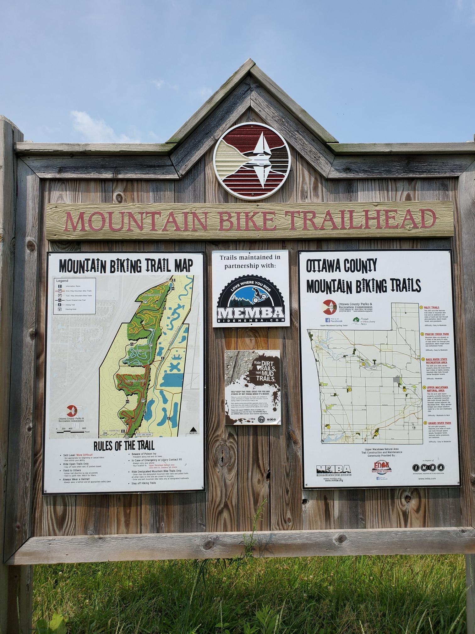 A wooden mountain bike trailhead sign with maps under a bright sky.