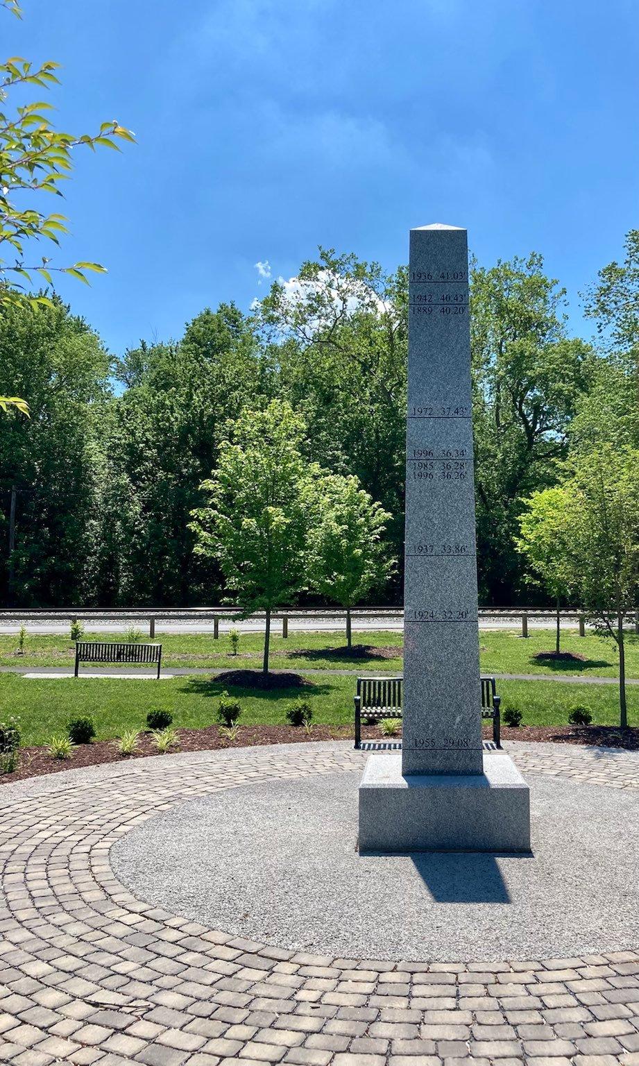 A tall gray stone obelisk stands on a paved circle on a sunny day.