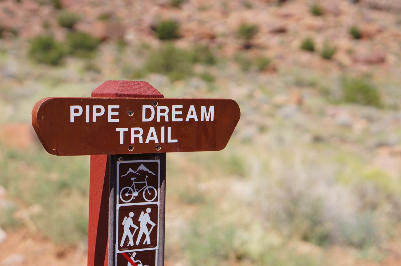 Brown trail sign with hiking and biking icons in a desert landscape. - Pipe Dream – Best of Moab MTB Trails