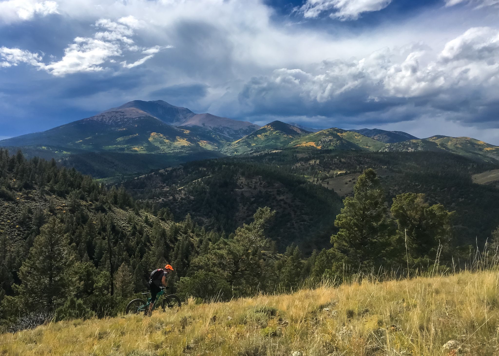 Mountain biker on a grassy slope with dramatic mountains and cloudy sky. - Silver Creek - Rainbow Trail Loop