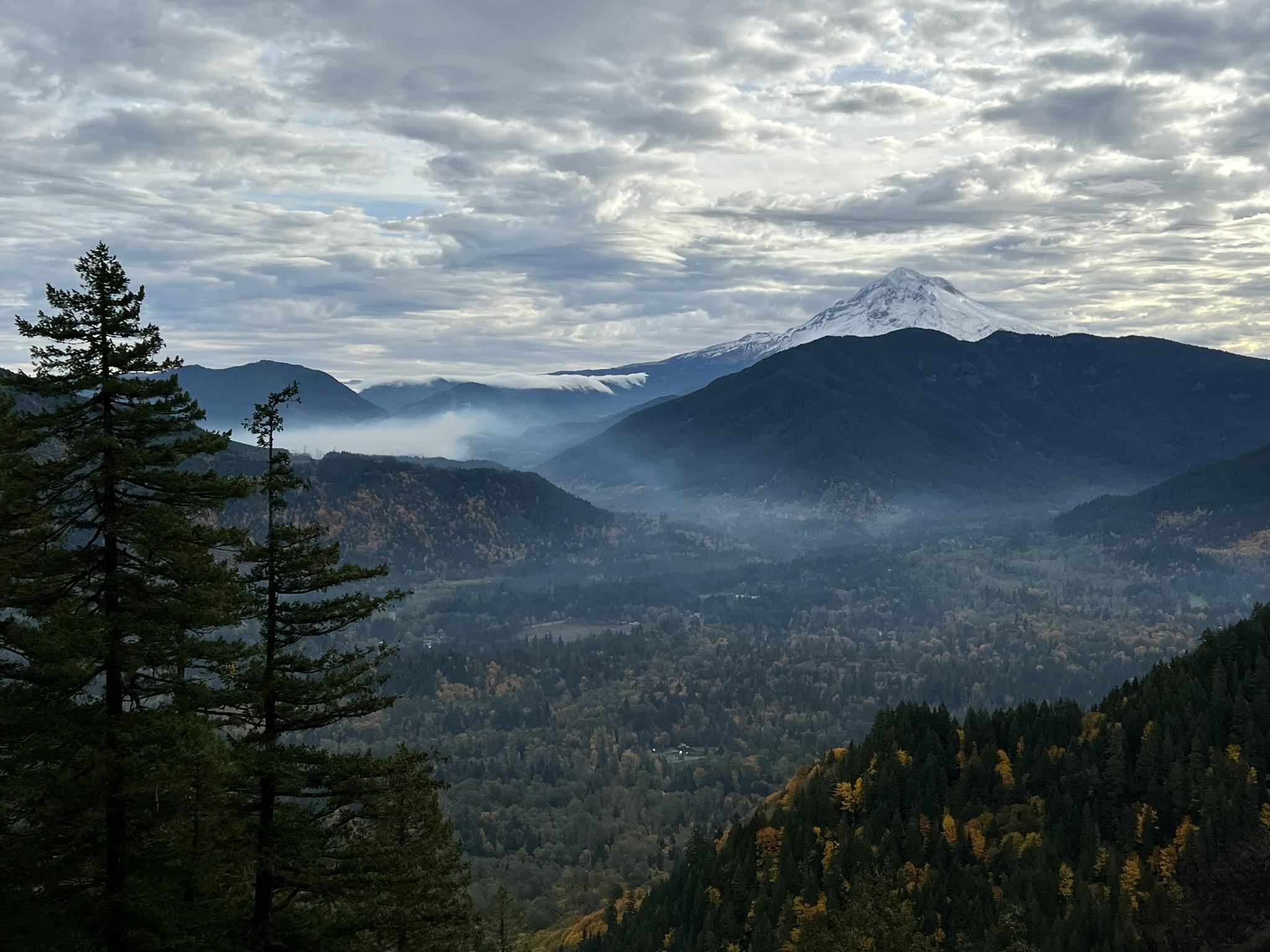 A snow-capped mountain rises above a misty, forested valley under clouds.