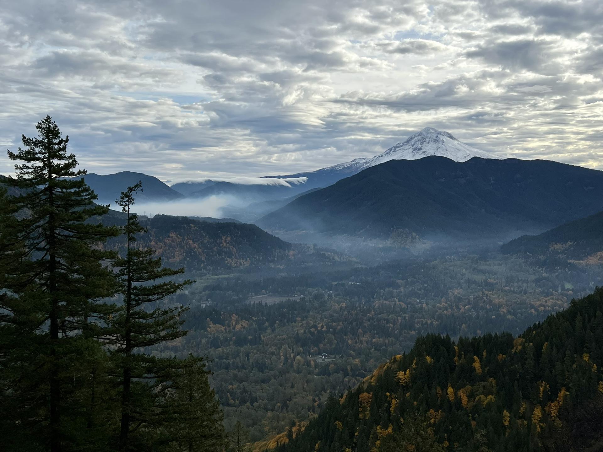 A snow-capped mountain rises above a misty, forested valley under clouds.