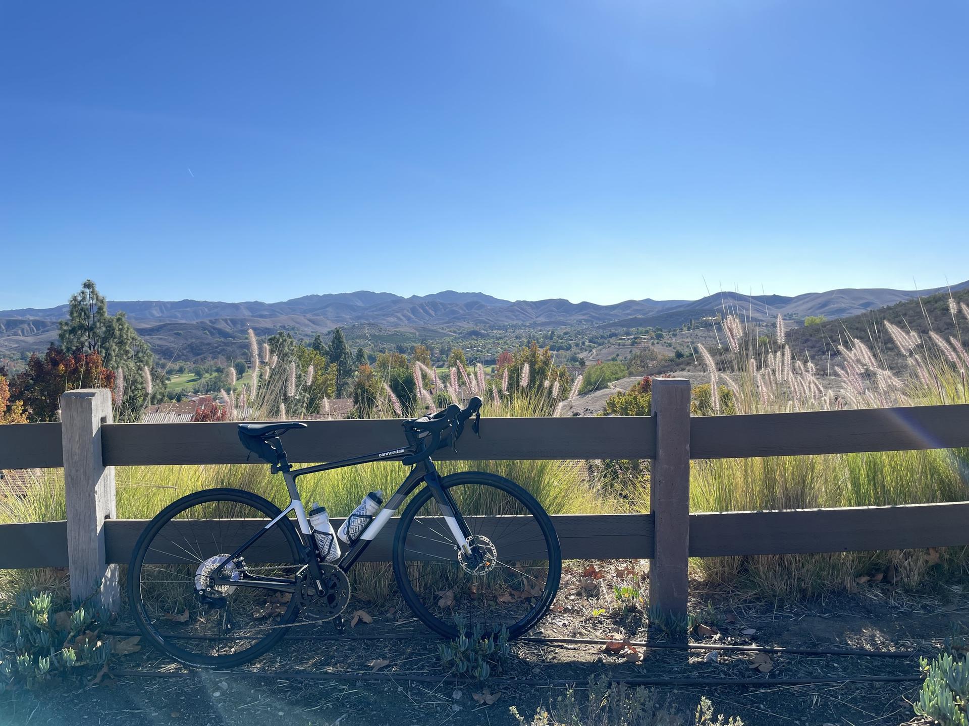 A bicycle leans on a fence overlooking sunny mountains and grassy hills.