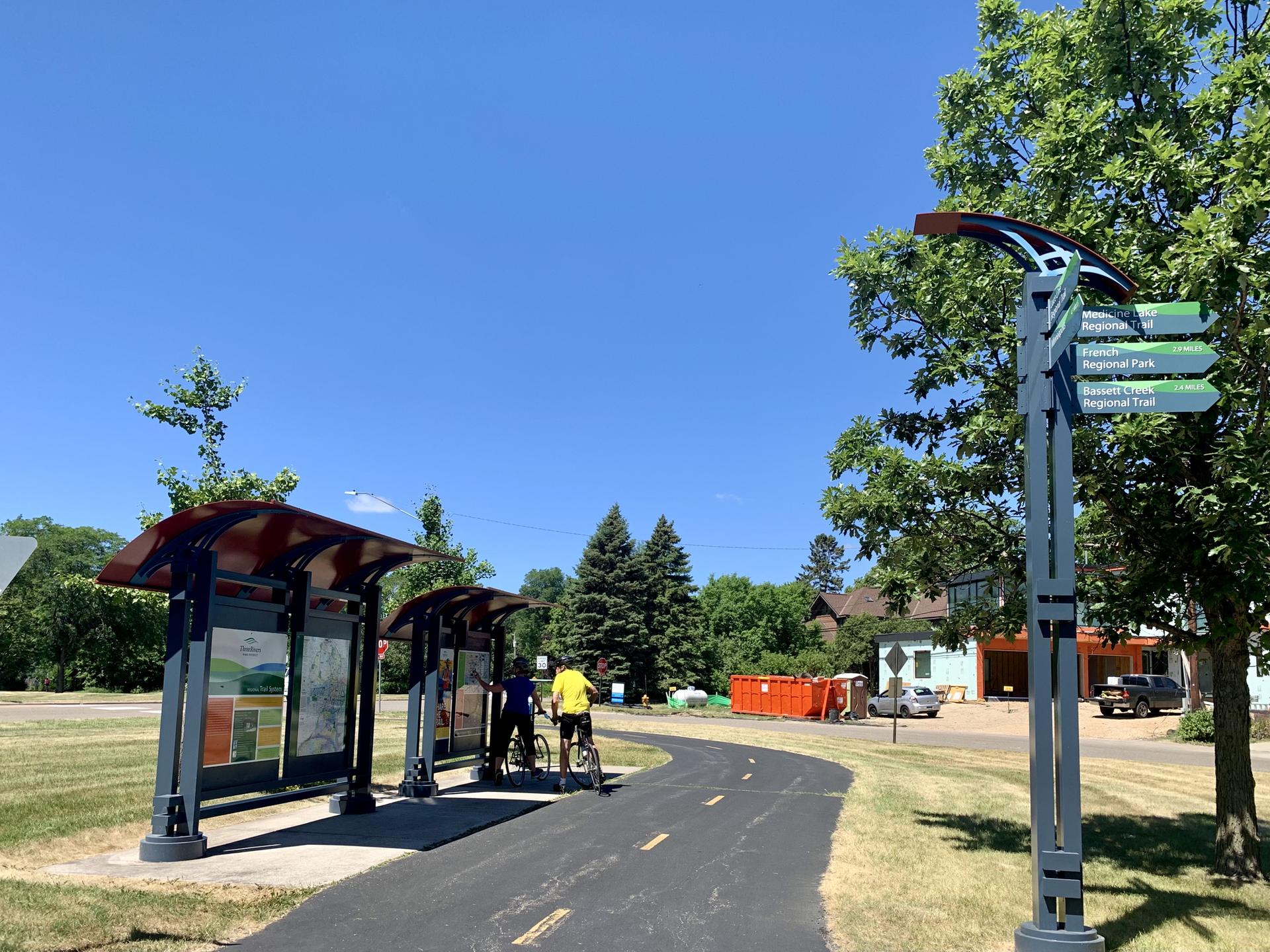 Two people with bikes near path shelters and a signpost on a sunny day.