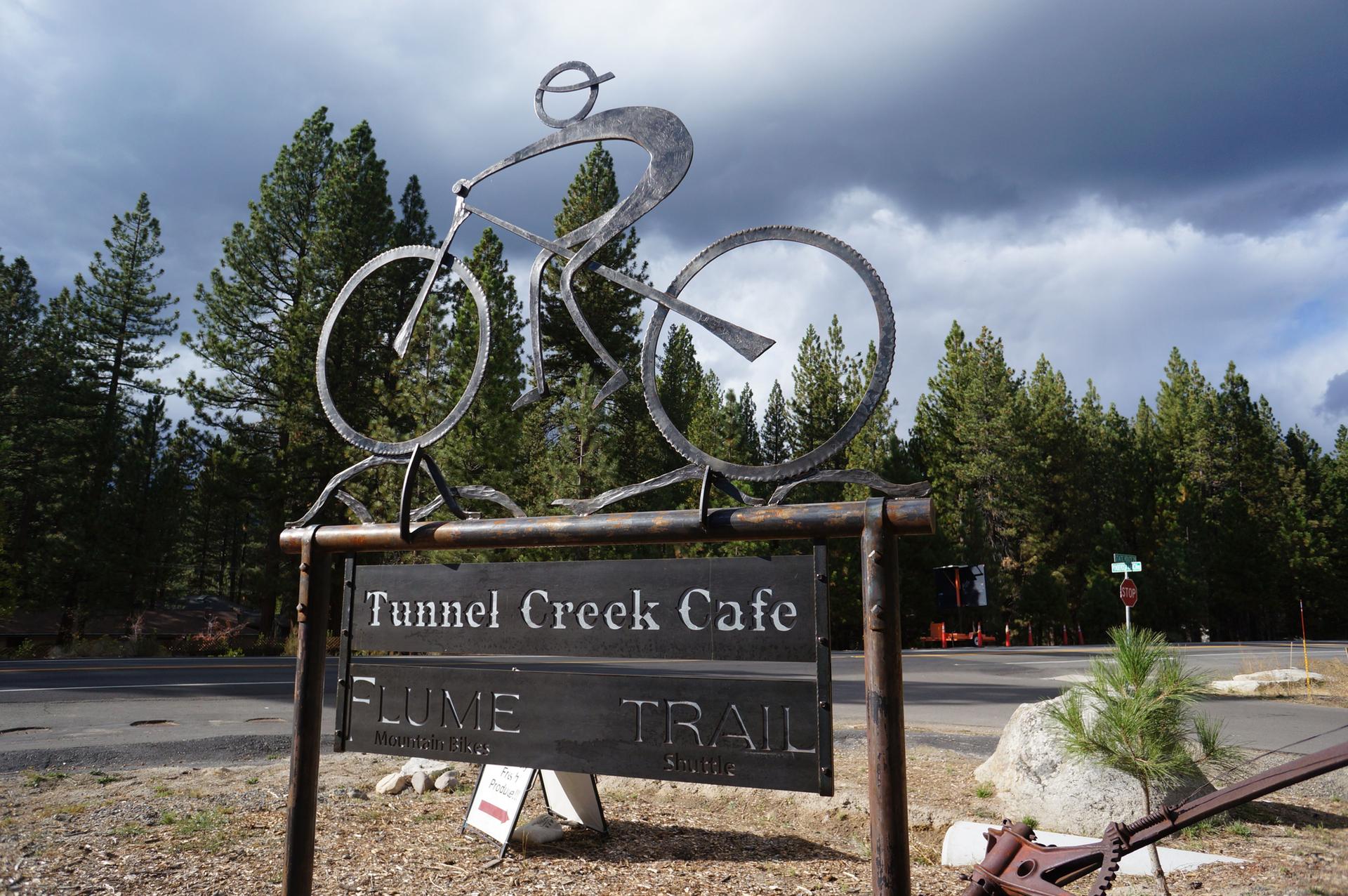 Metal cyclist sculpture on a sign, with pine trees and cloudy sky.