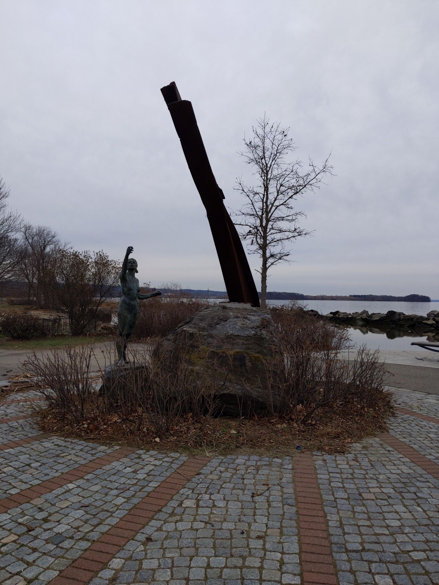 A bronze statue and dark sculpture near water, with bare trees and an overcast sky. - Croton Riverwalk