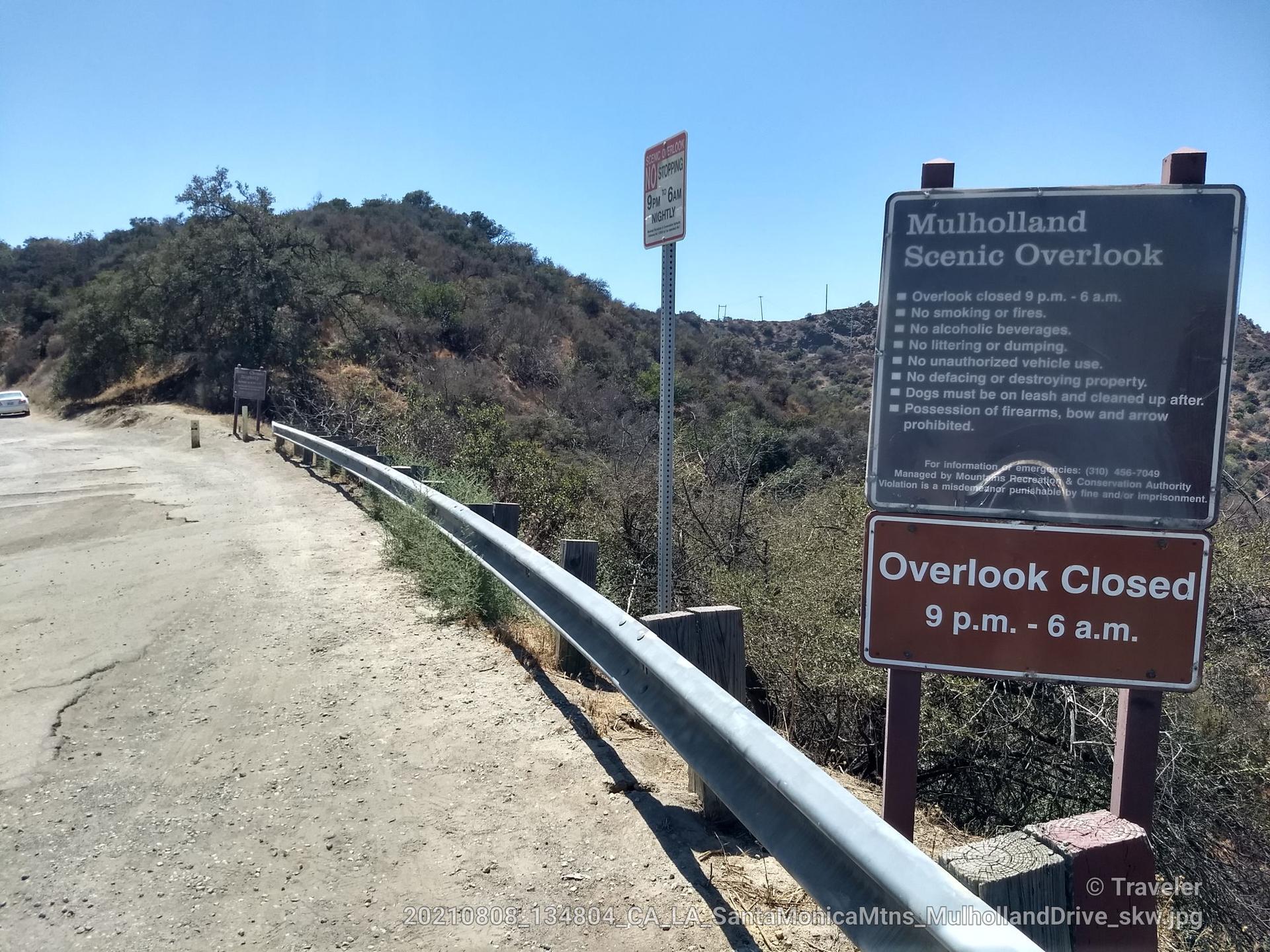 Roadside signs and guardrail on a sunny day with dry, hilly terrain.