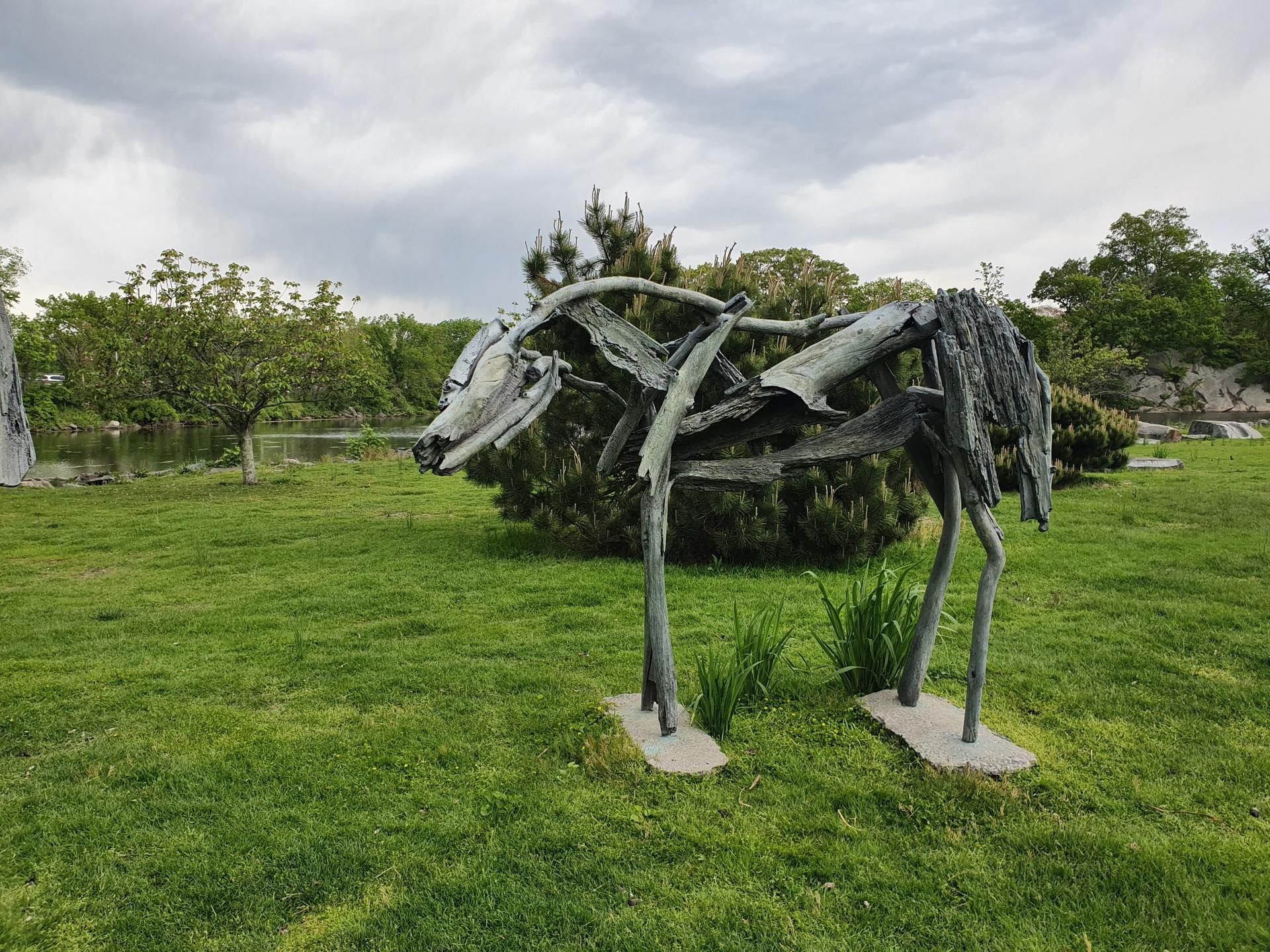 Wooden horse sculpture in a grassy park with a pond under an overcast sky.