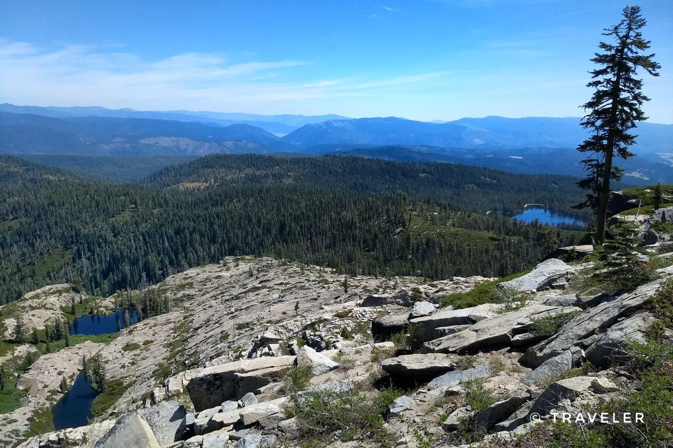 Expansive mountain vista with rocky terrain, dense forests, and multiple small lakes.