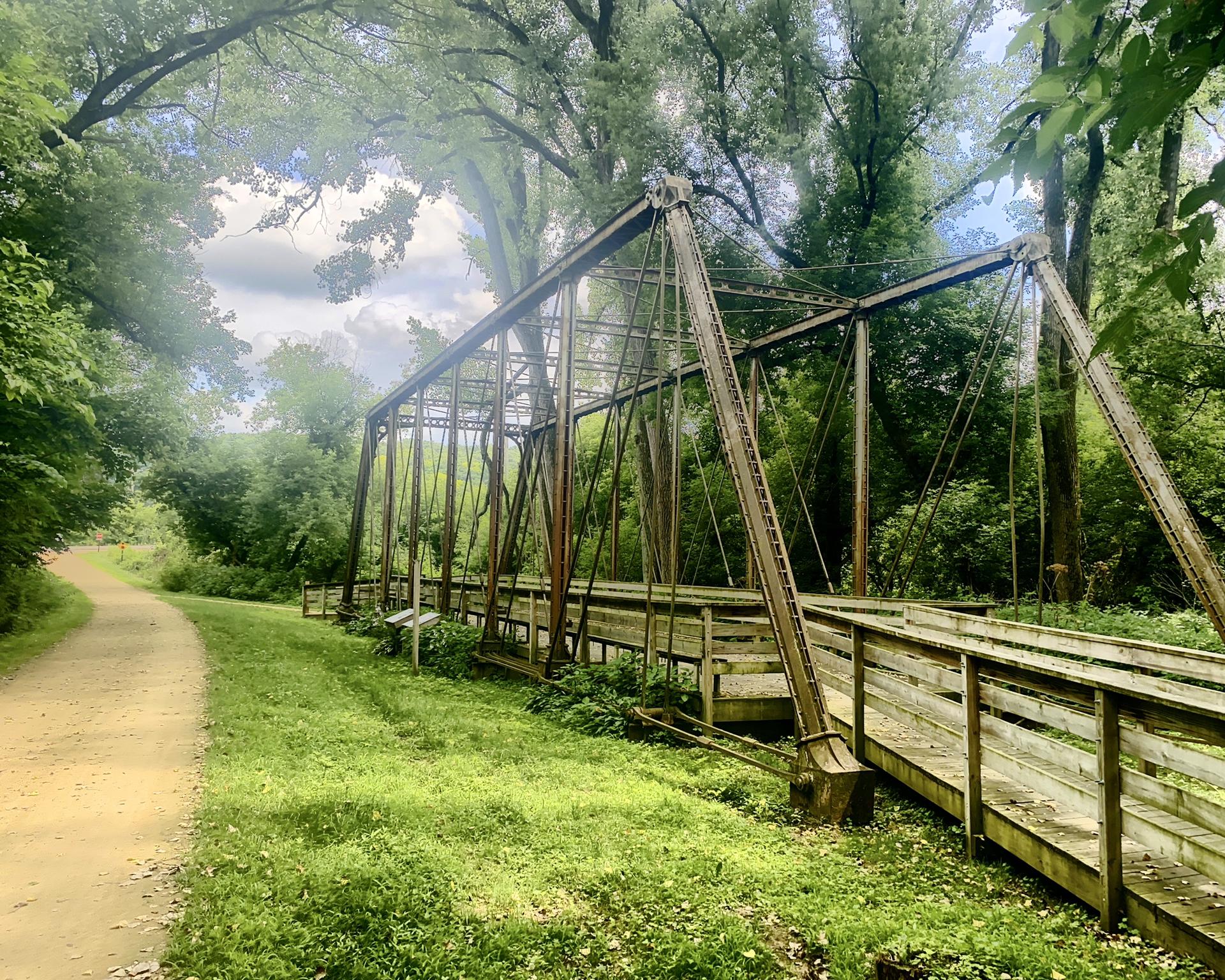 An old metal bridge spans a lush green forest beside a dirt path.