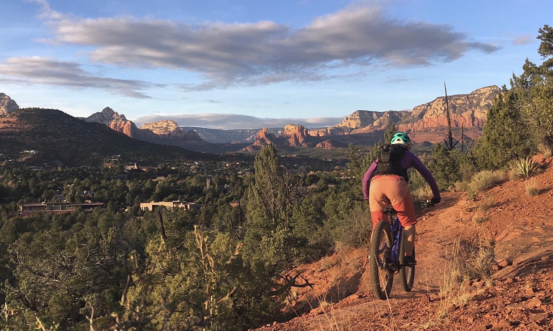 Person mountain biking on a red dirt trail with distant mountains at sunset.