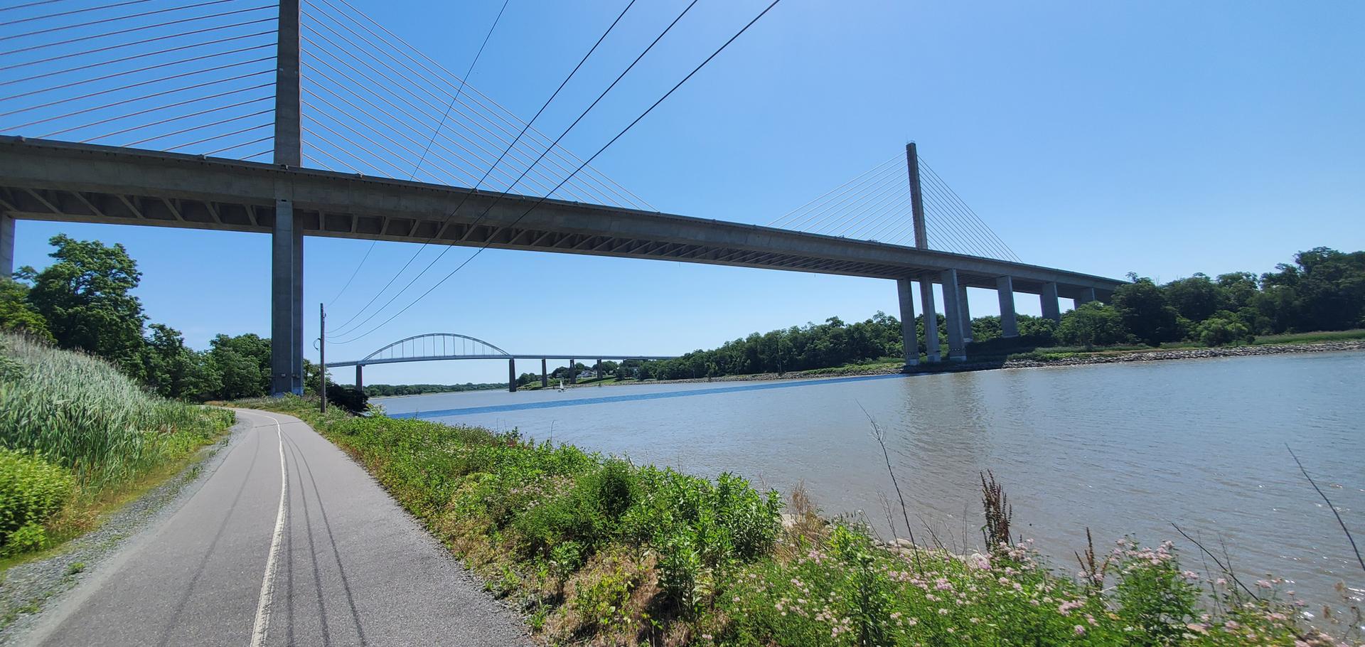 A paved path runs alongside a river with two large bridges overhead.