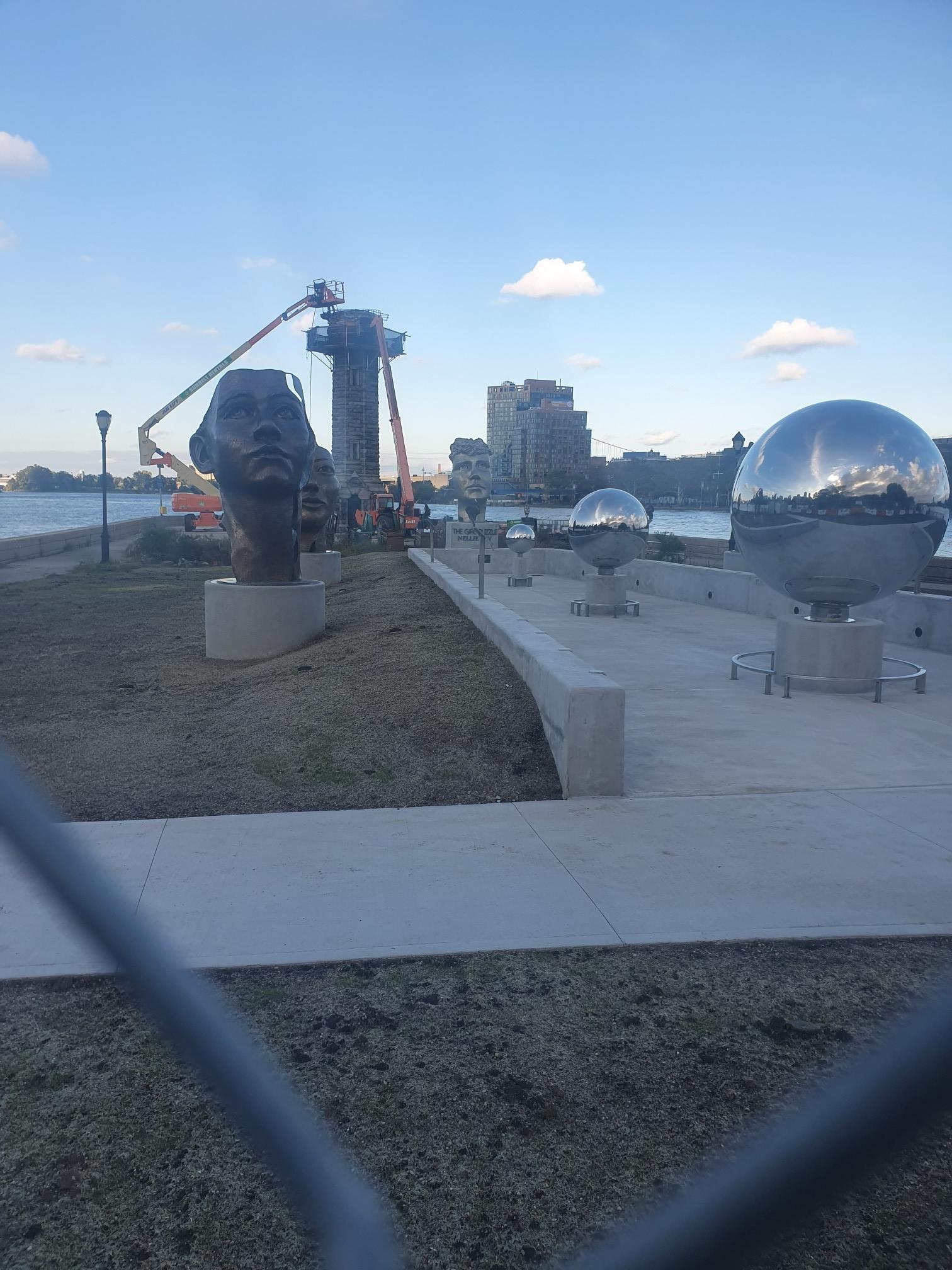 Outdoor sculpture park with large head and spheres, city construction in background.