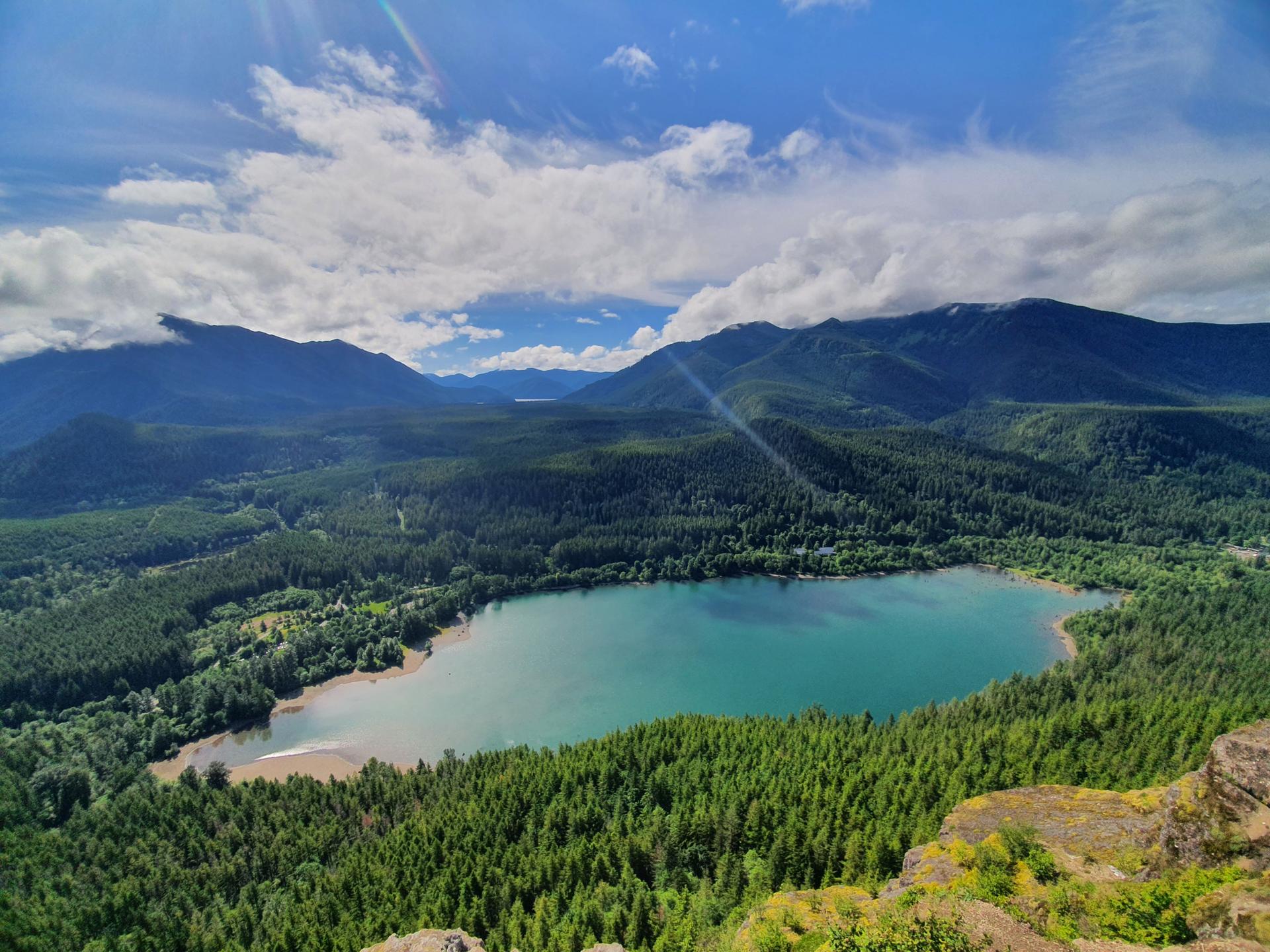 Bright sunny view of a turquoise lake surrounded by dense green forests and mountains.