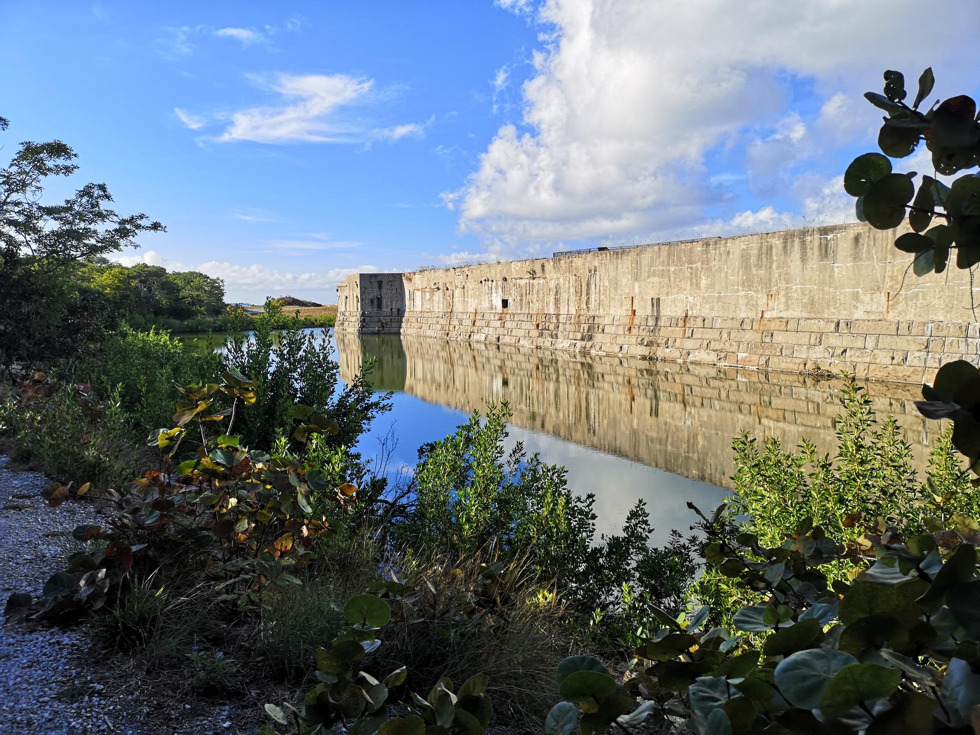 Large stone wall reflecting in calm water, surrounded by green foliage.