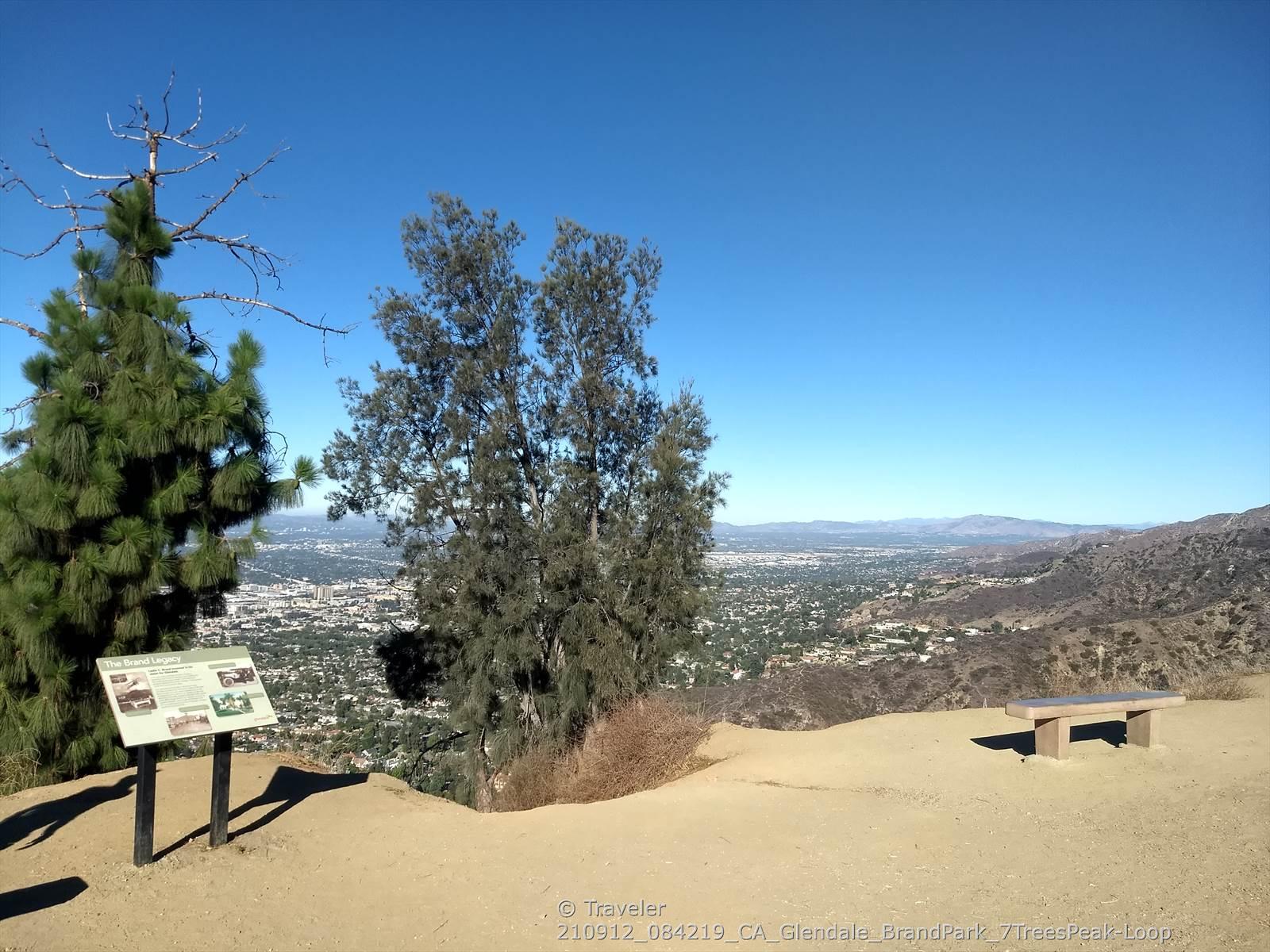 Overlook with trees, bench, and informational sign, city below, clear sky.