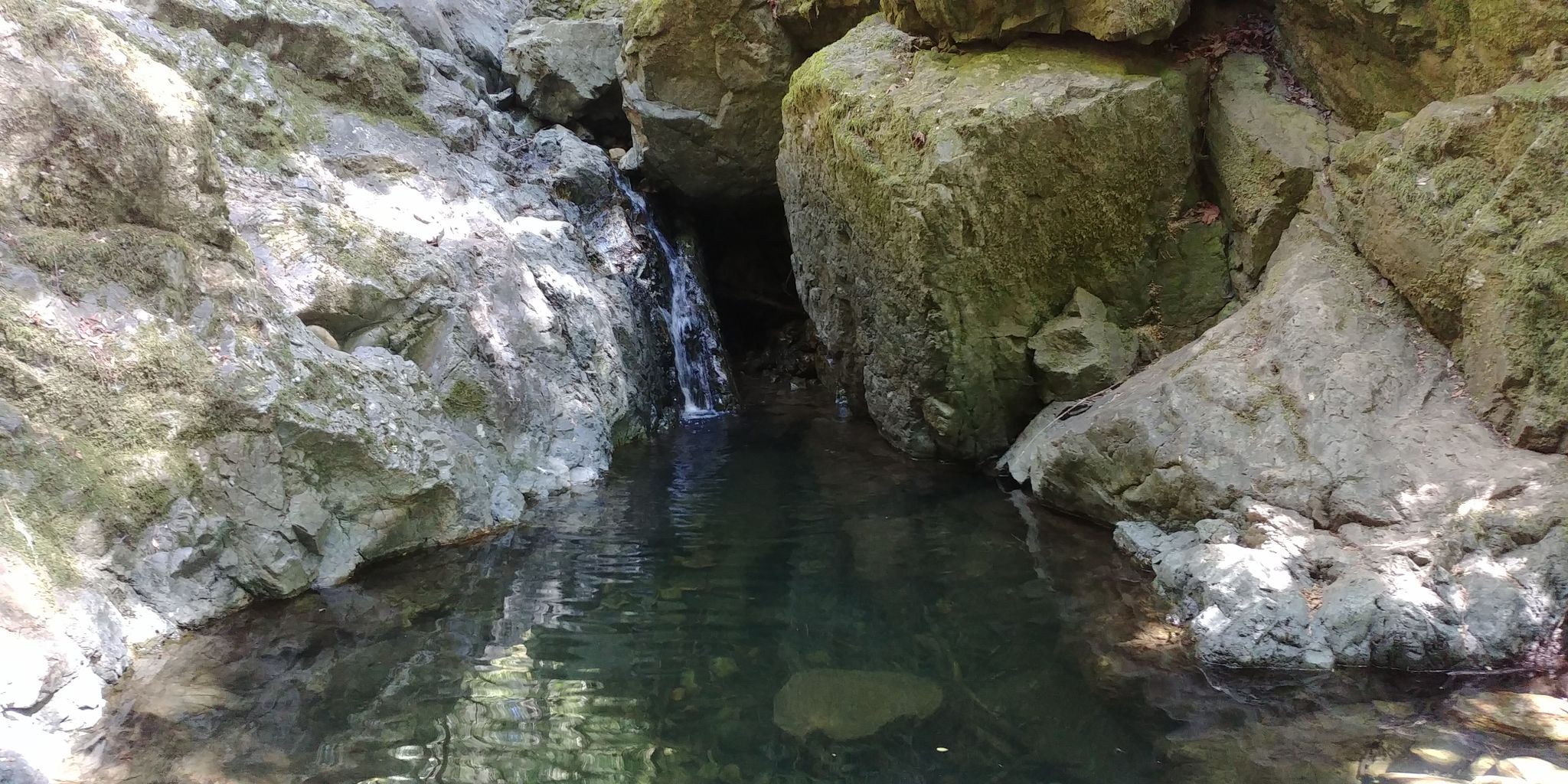 Dark pool of water surrounded by large rocks, with a small waterfall. - Cataract Falls Loop