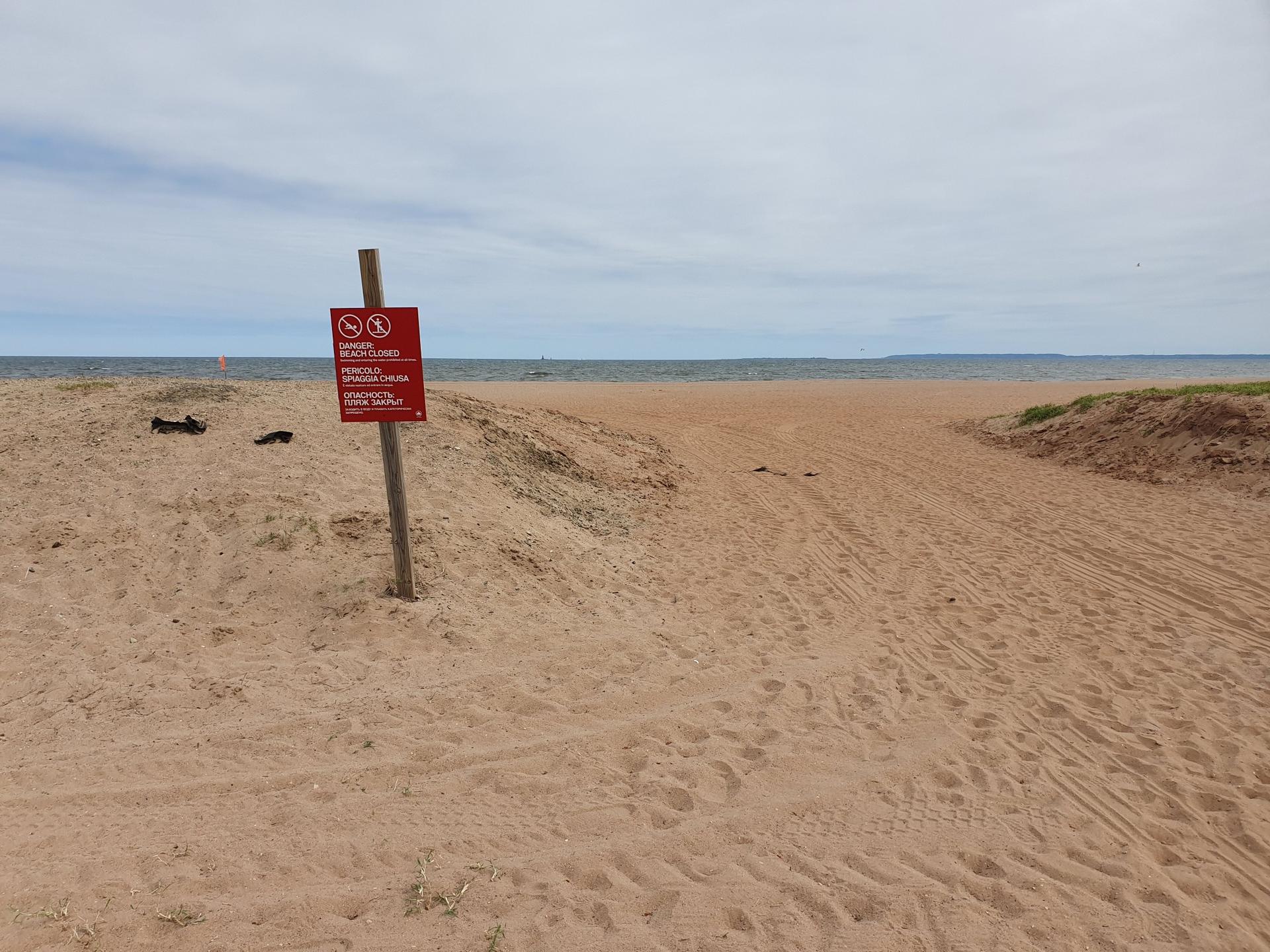 Sandy beach with a red warning sign, ocean in distance under cloudy sky.