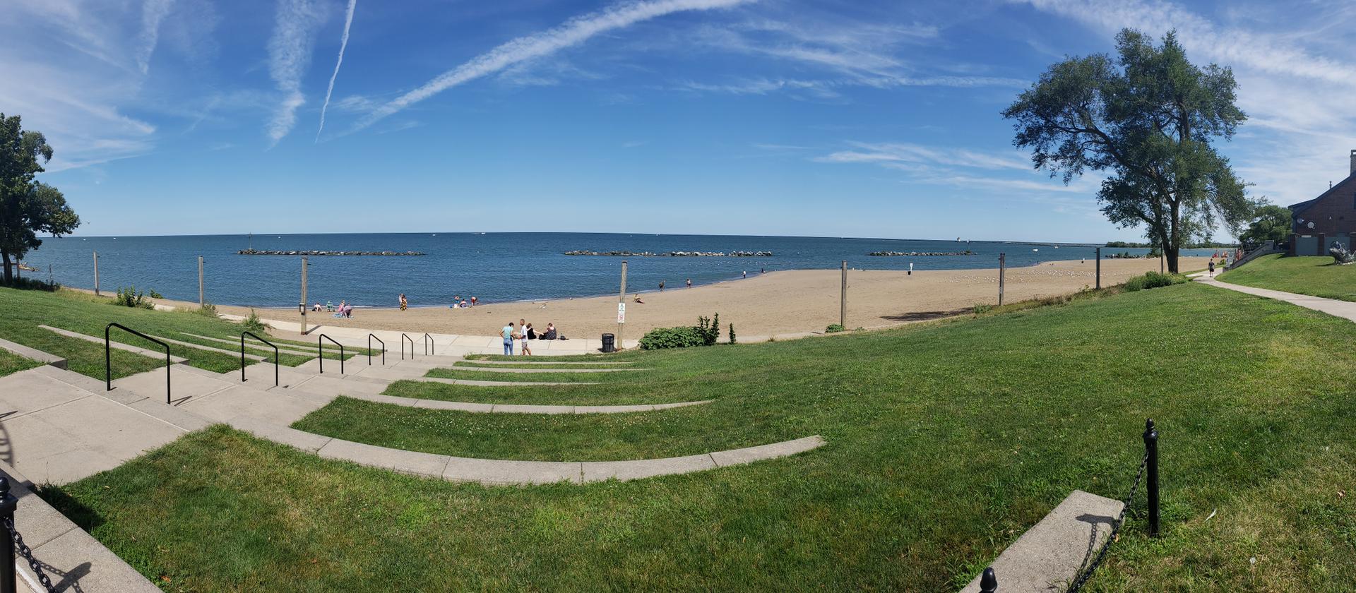 Panoramic sunny beach with people, blue ocean, and grassy steps.