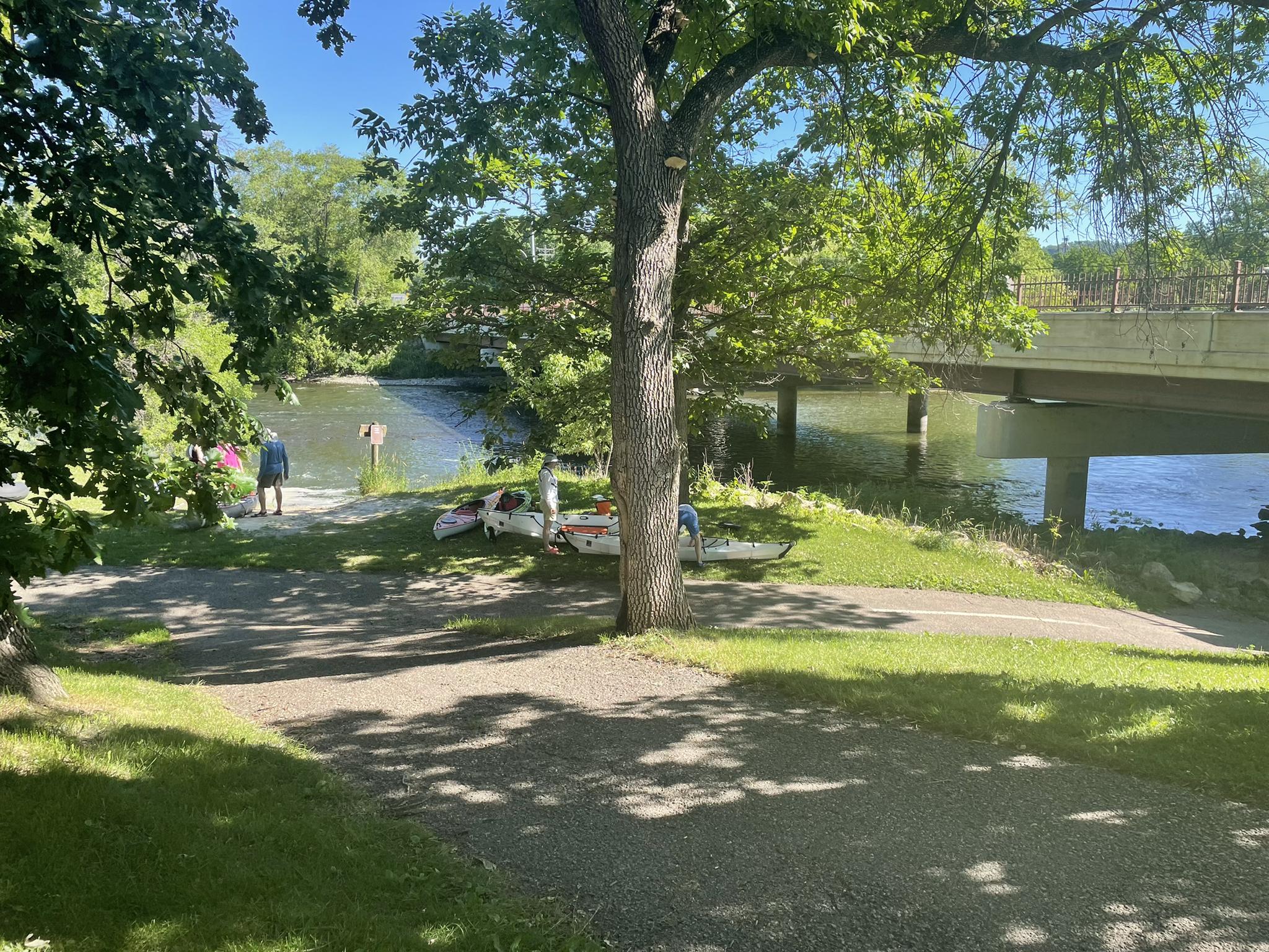 People and kayaks by a river under a bridge on a sunny day.