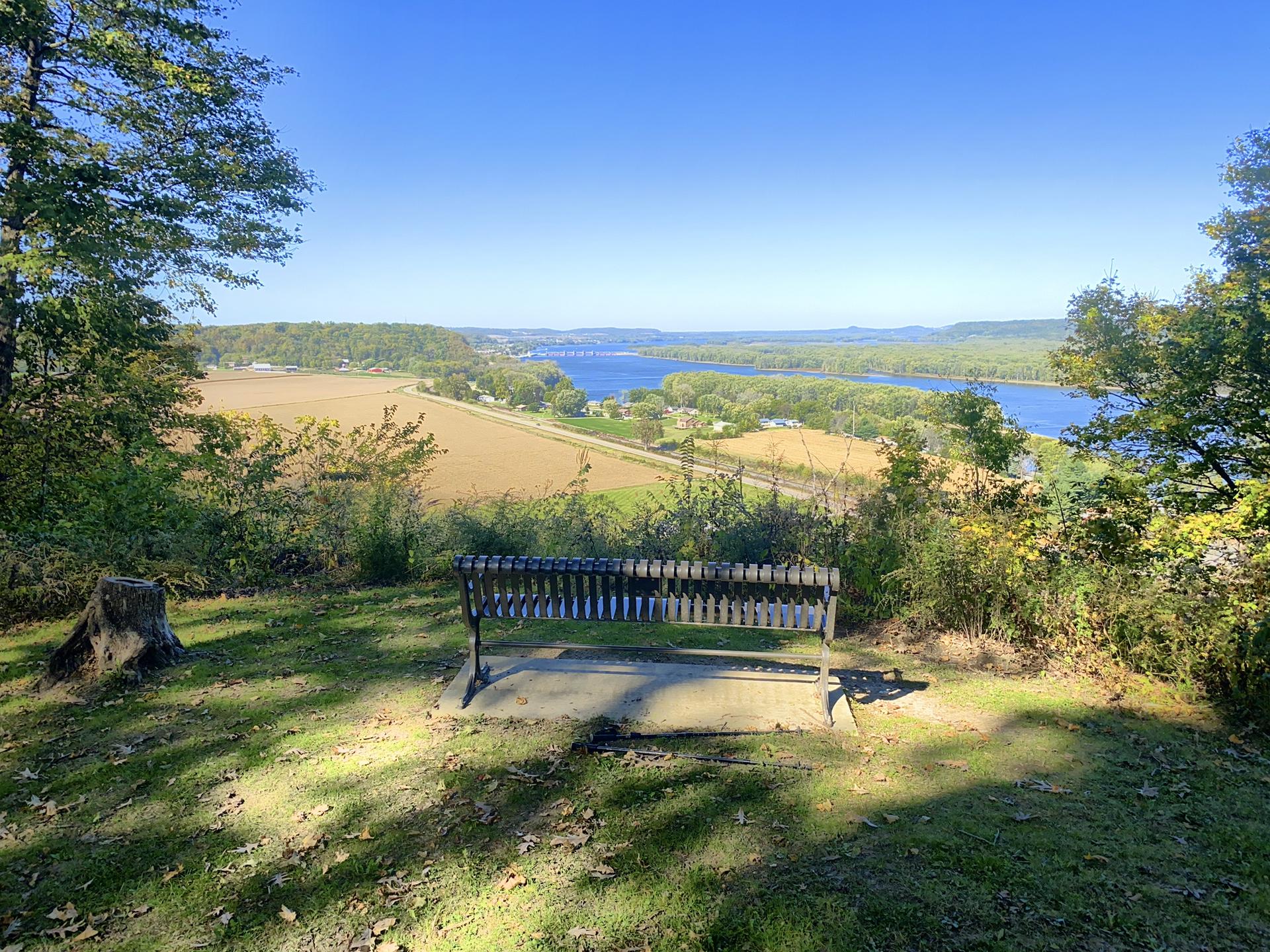 Empty park bench overlooks a sunny river valley with fields and trees.