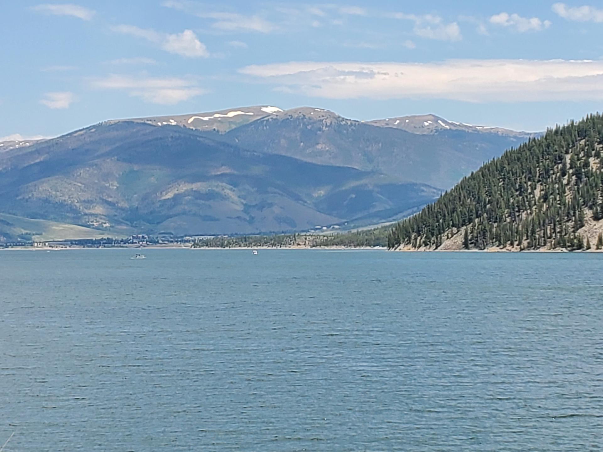 Blue lake, forested hills, distant snow-capped mountains, and small boats under cloudy sky.