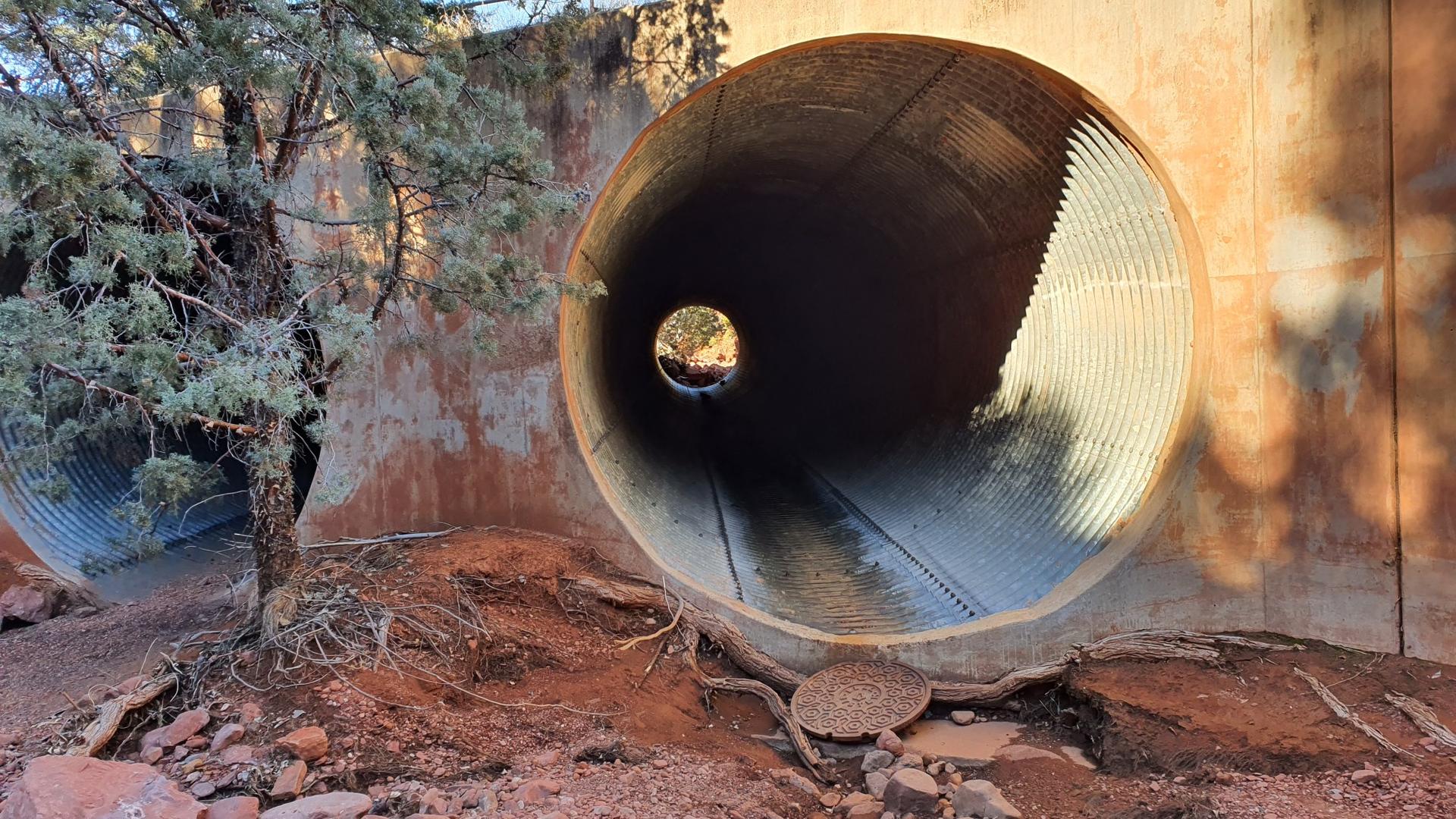 Large metal culvert tunnel with light at the end, surrounded by red earth.