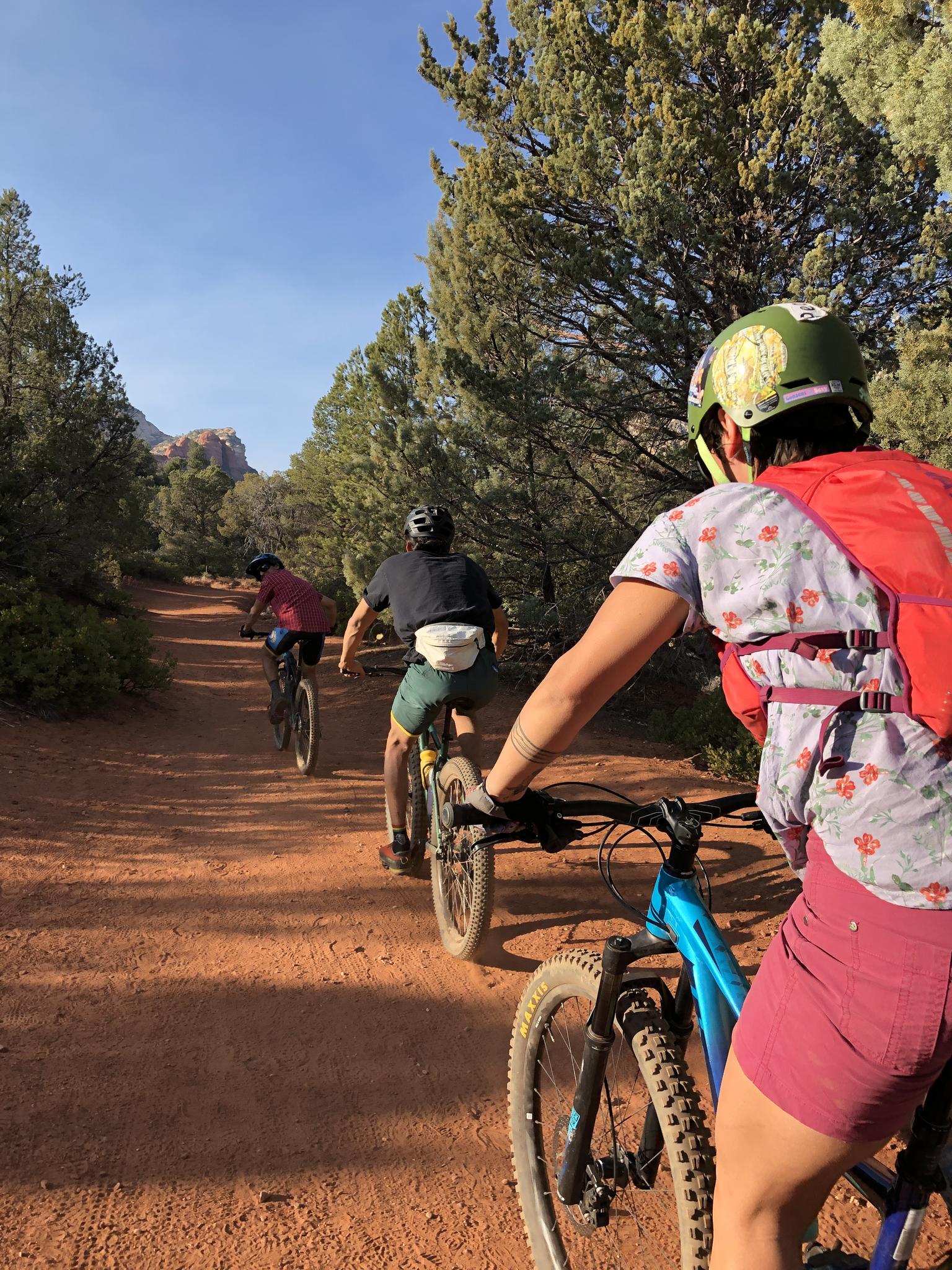 Three mountain bikers ride on a red dirt trail through green trees.
