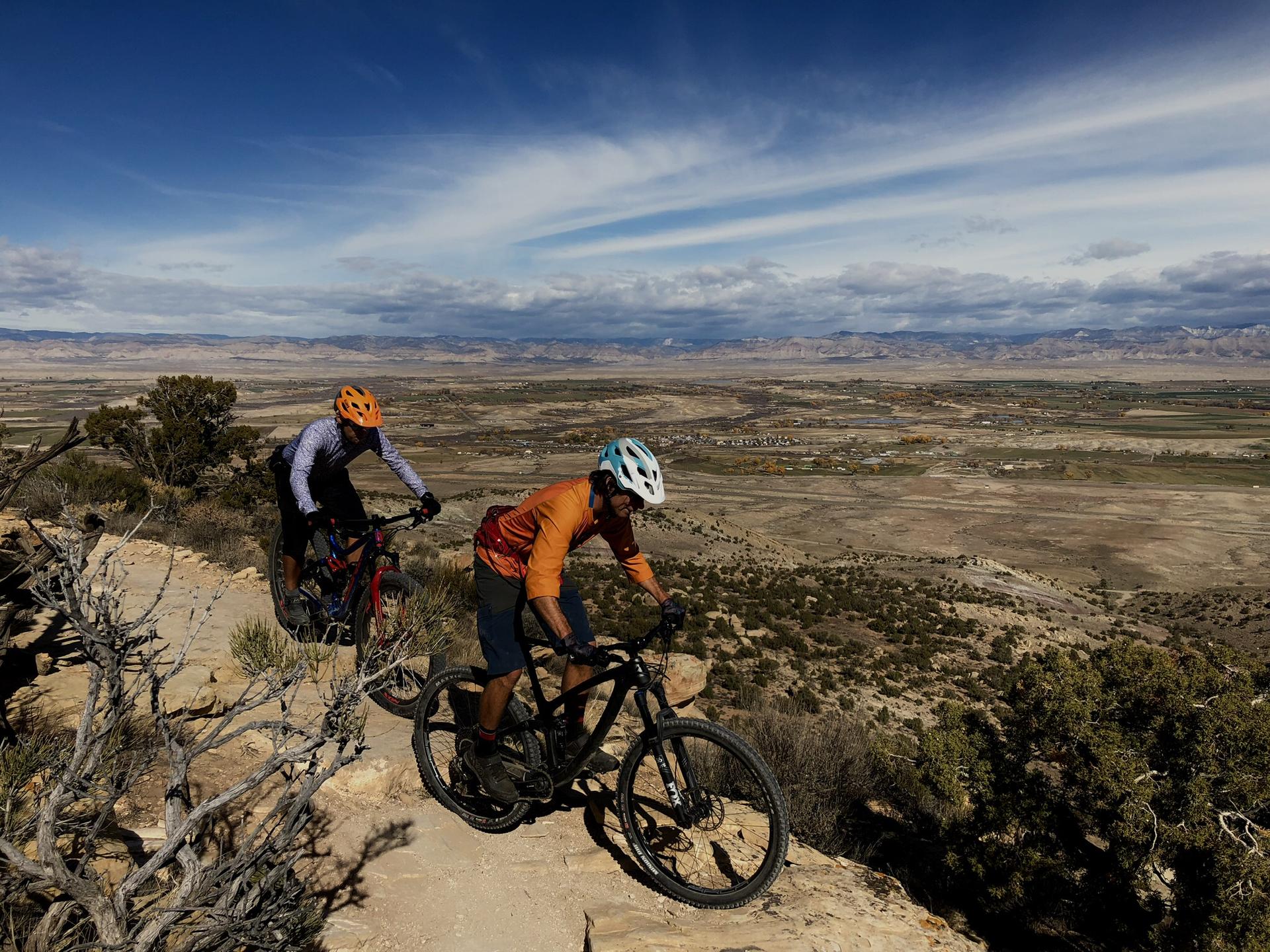 Two mountain bikers on a rocky trail overlooking a vast arid landscape.