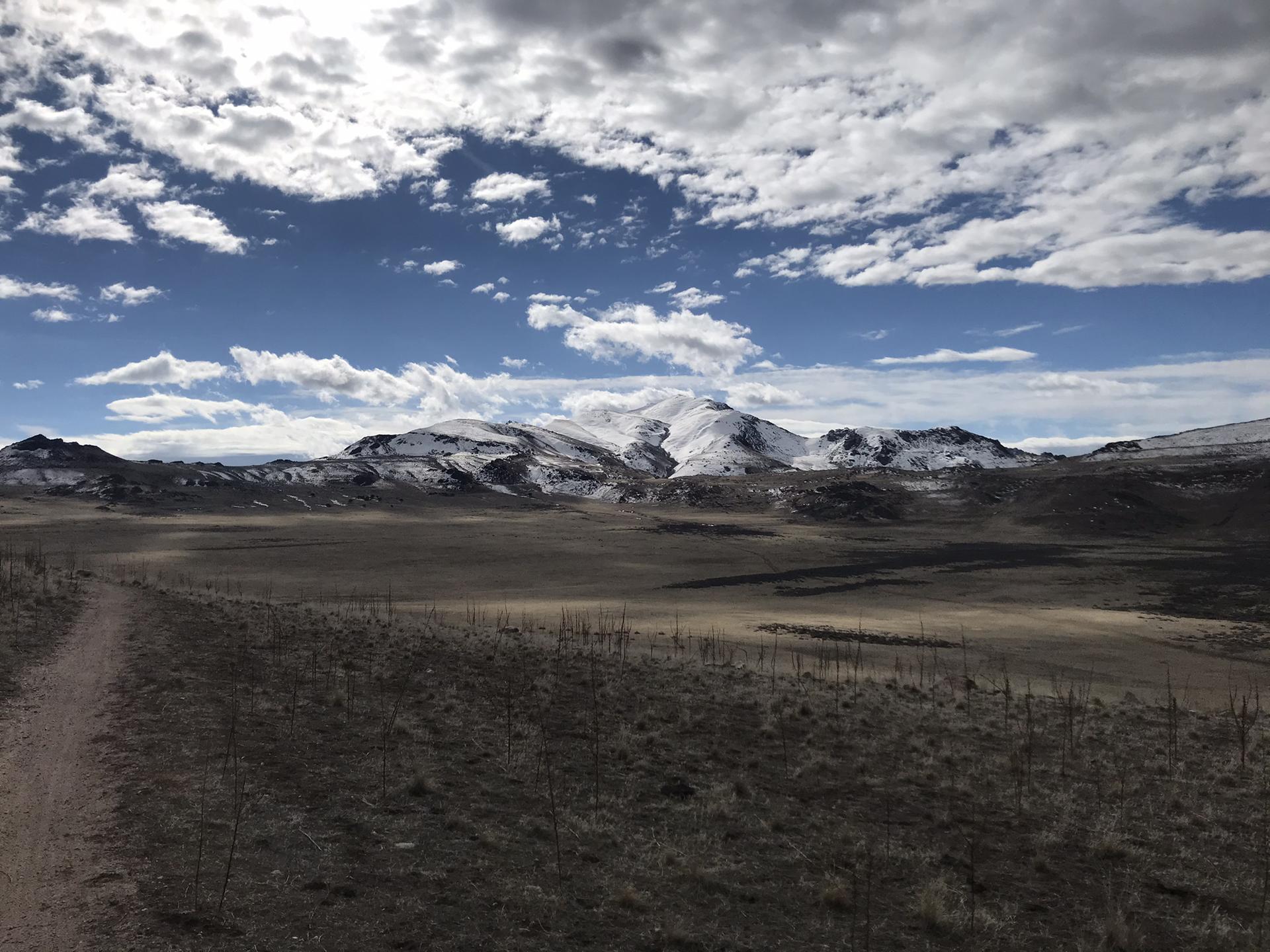 Dirt path leads towards snow-capped mountains under a dramatic cloudy sky.