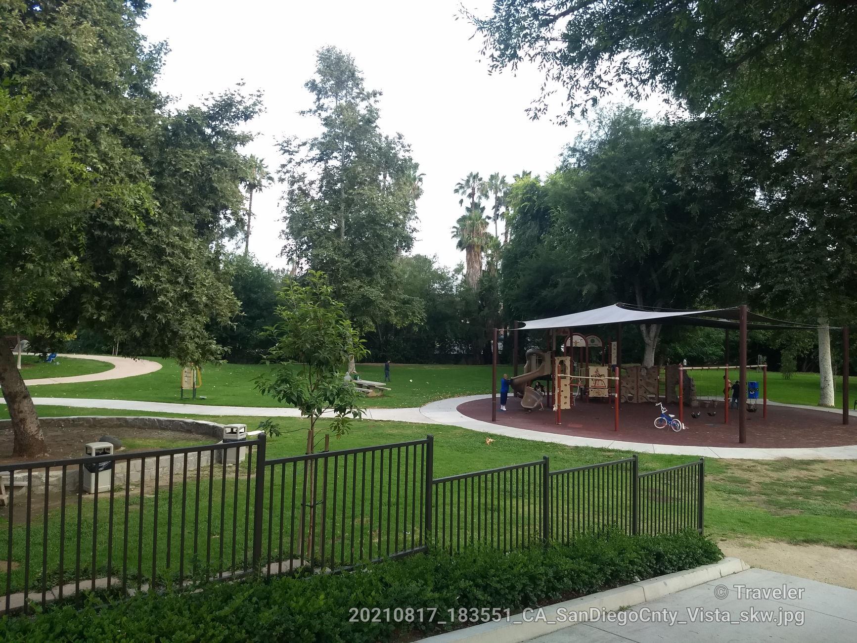 A park playground with a canopy, green grass, and tall trees on an overcast day.