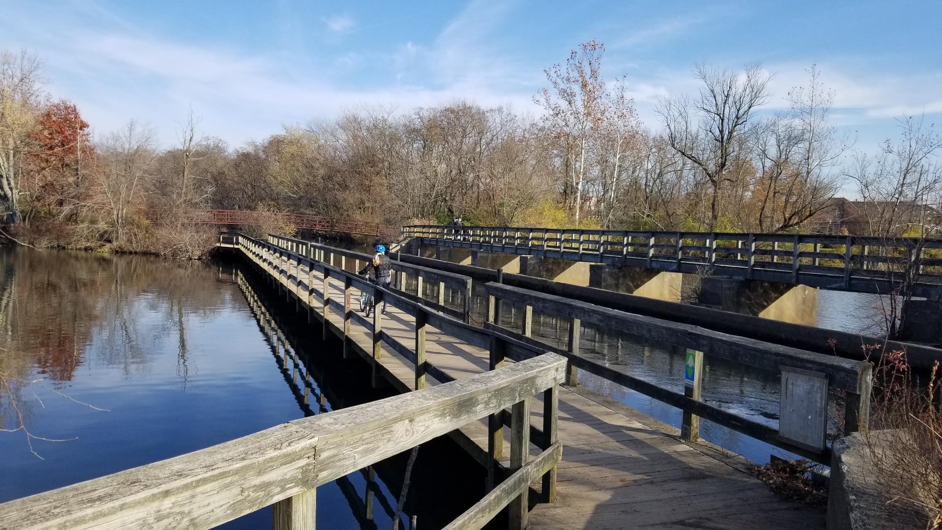 A person bikes across wooden bridges over calm water on a sunny day.