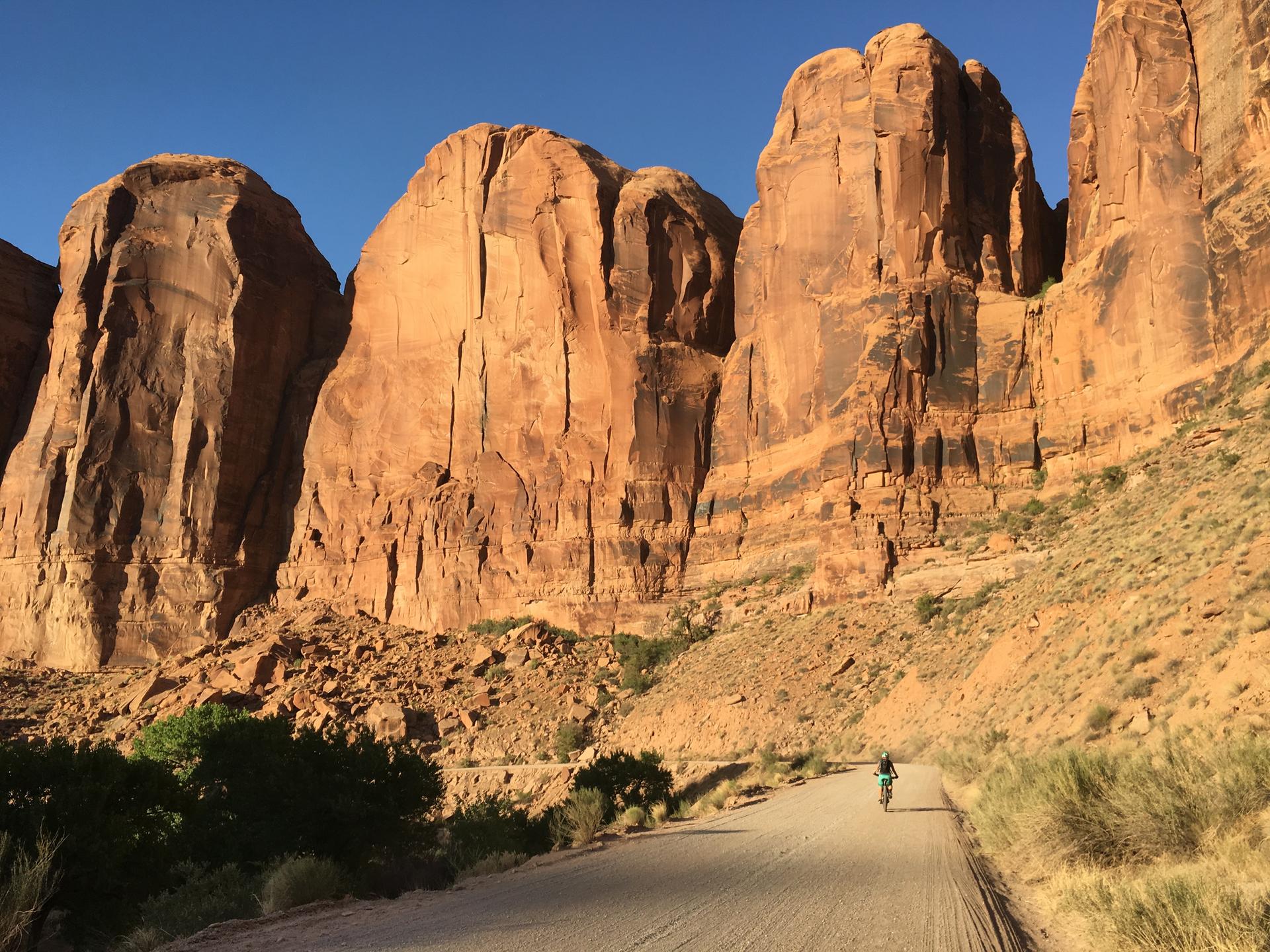 Distant mountain biker on a dirt road flanked by massive red rock formations.