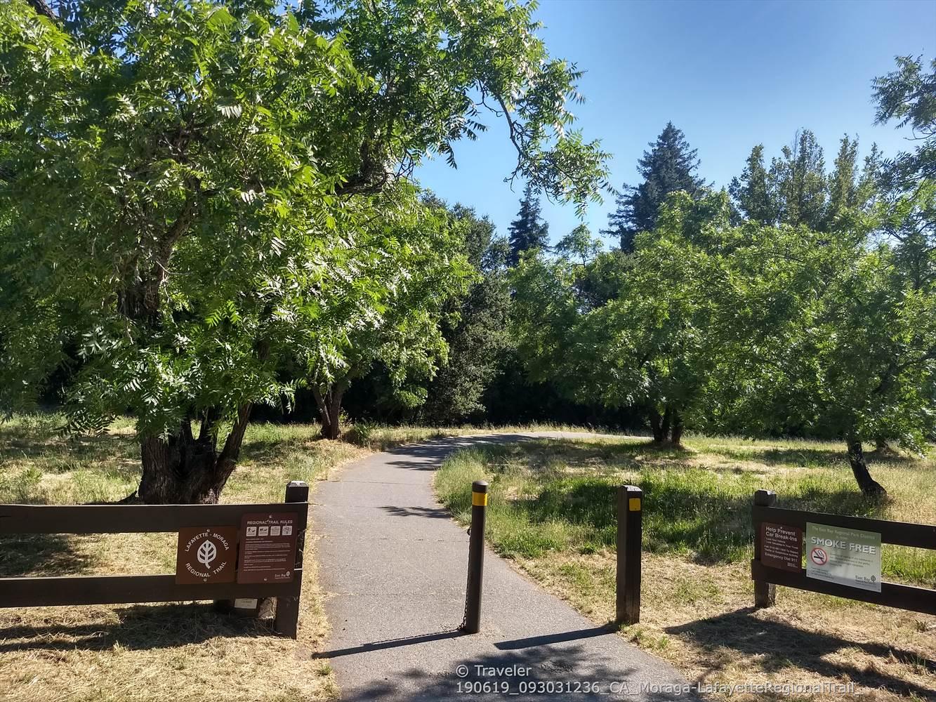 Paved path entrance with wooden fences, surrounded by green trees and dry grass.