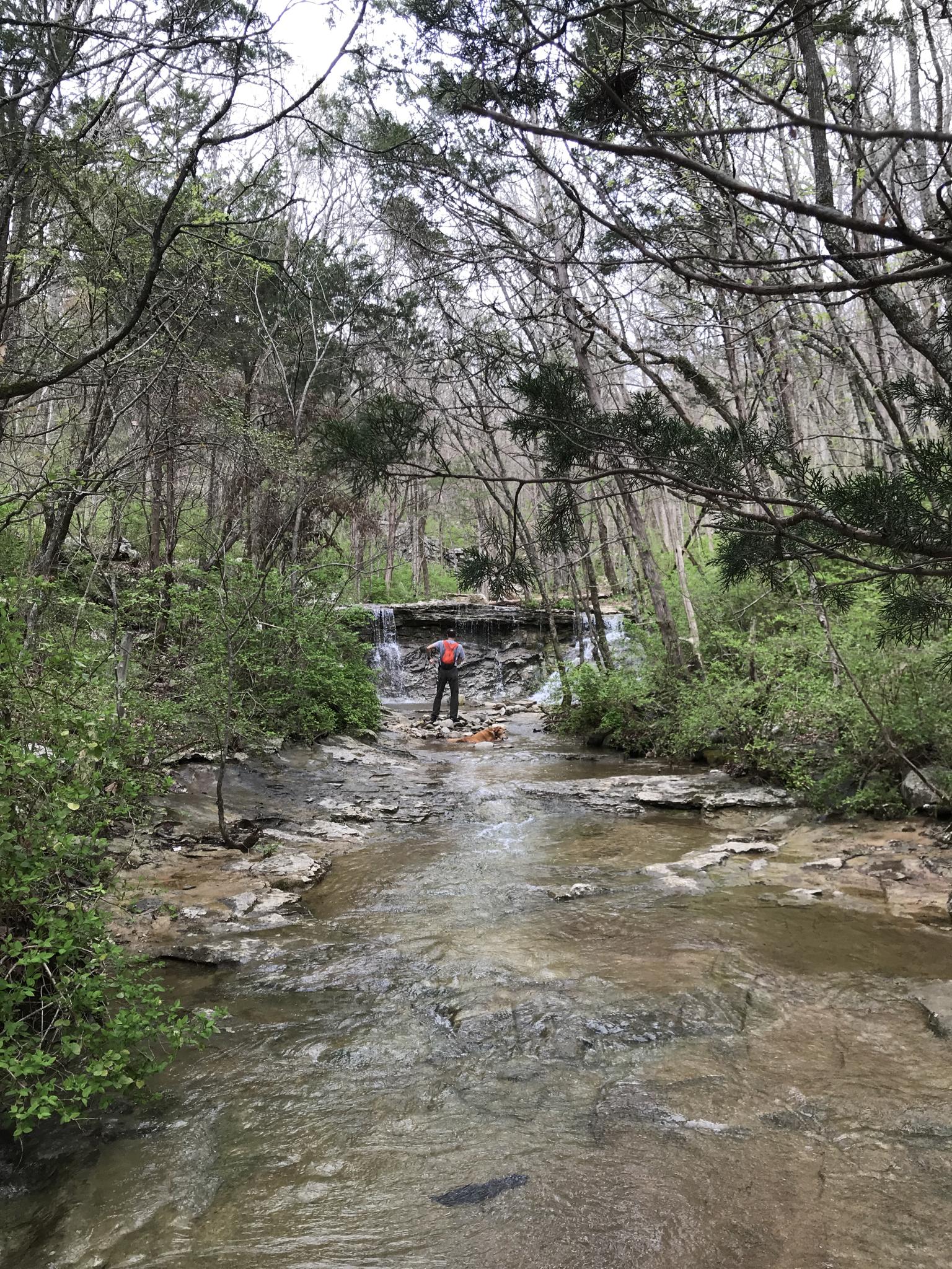 Person in red jacket standing in a rocky stream before a small waterfall.