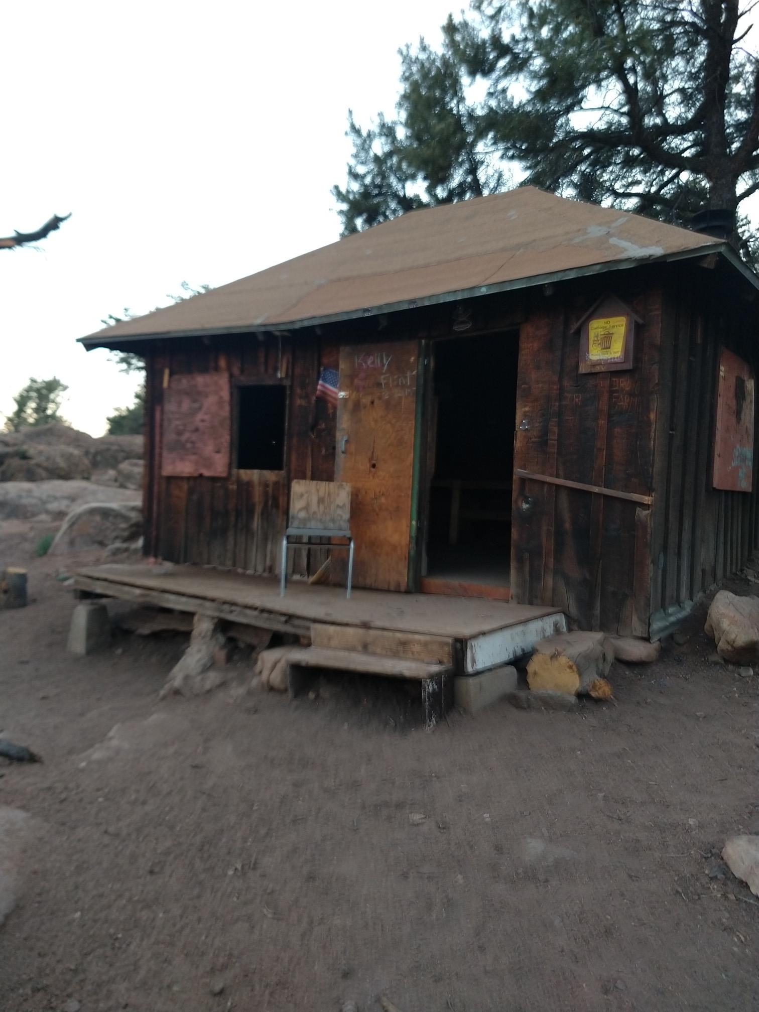 A rustic wooden cabin with a porch and chair, surrounded by dirt and trees.