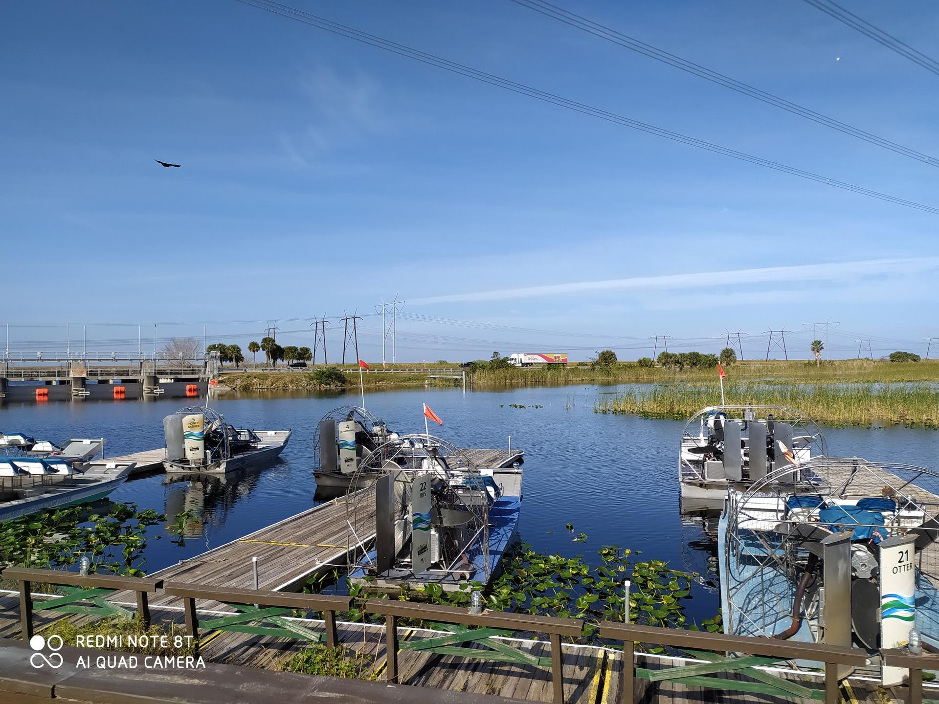Several airboats are docked at a wooden pier on a sunny day.