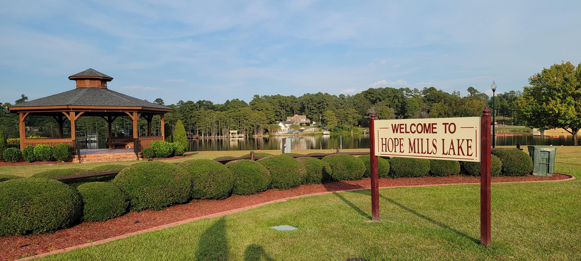 A wooden gazebo and sign by a calm lake with green bushes under a clear sky.