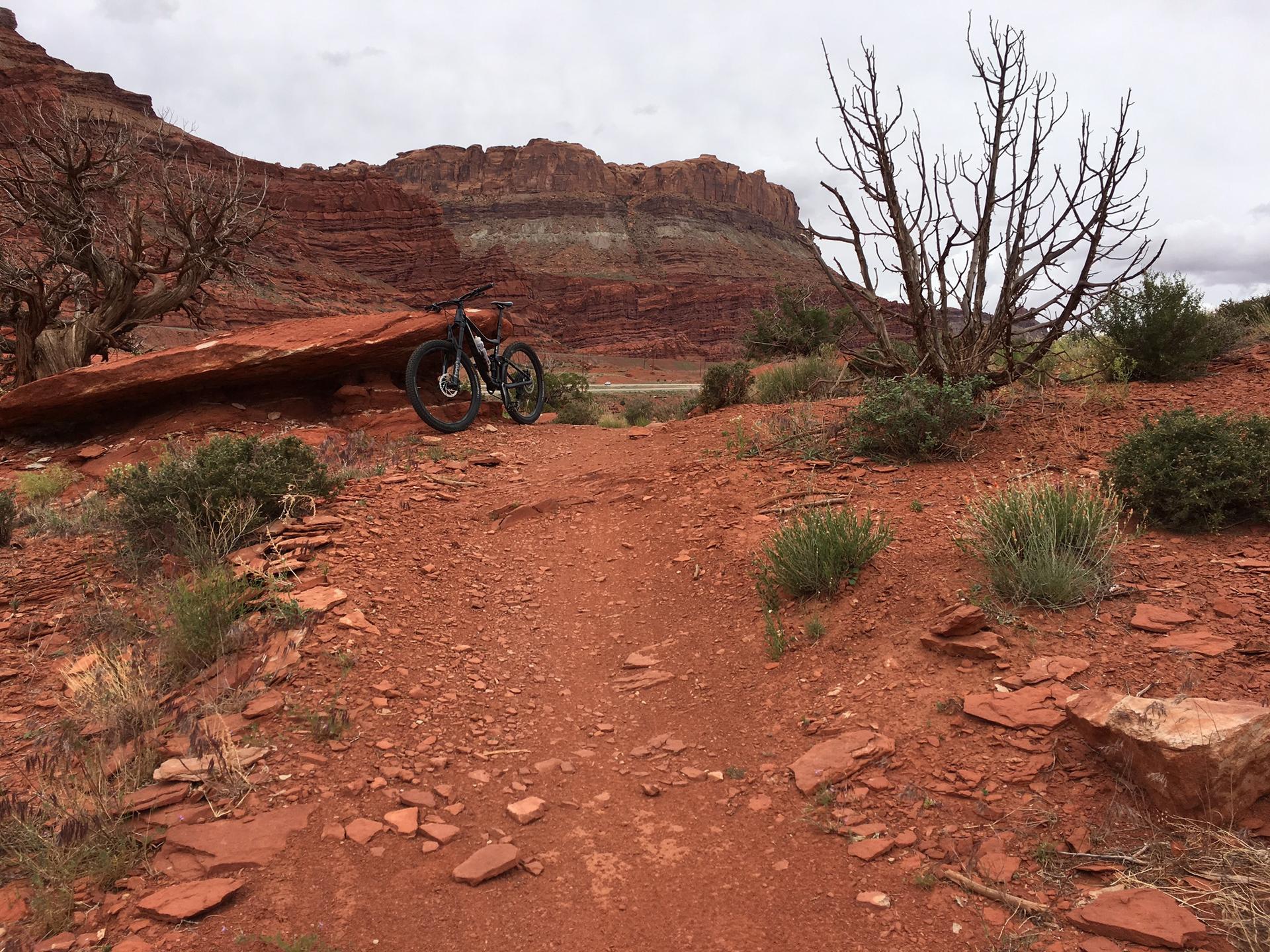 A mountain bike rests on a red dirt trail amidst sparse trees and cliffs.