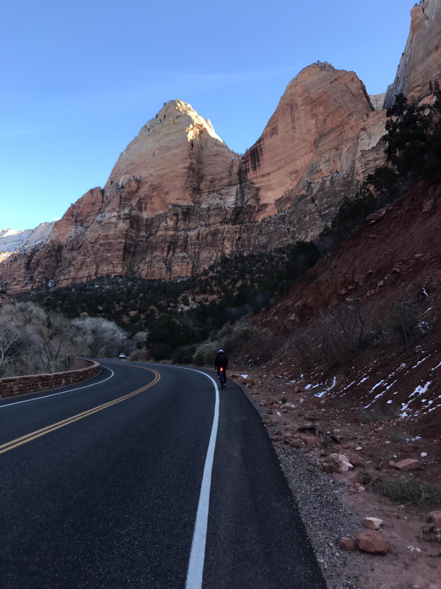 A person cycles on a winding road beside sunlit mountains with clear skies.