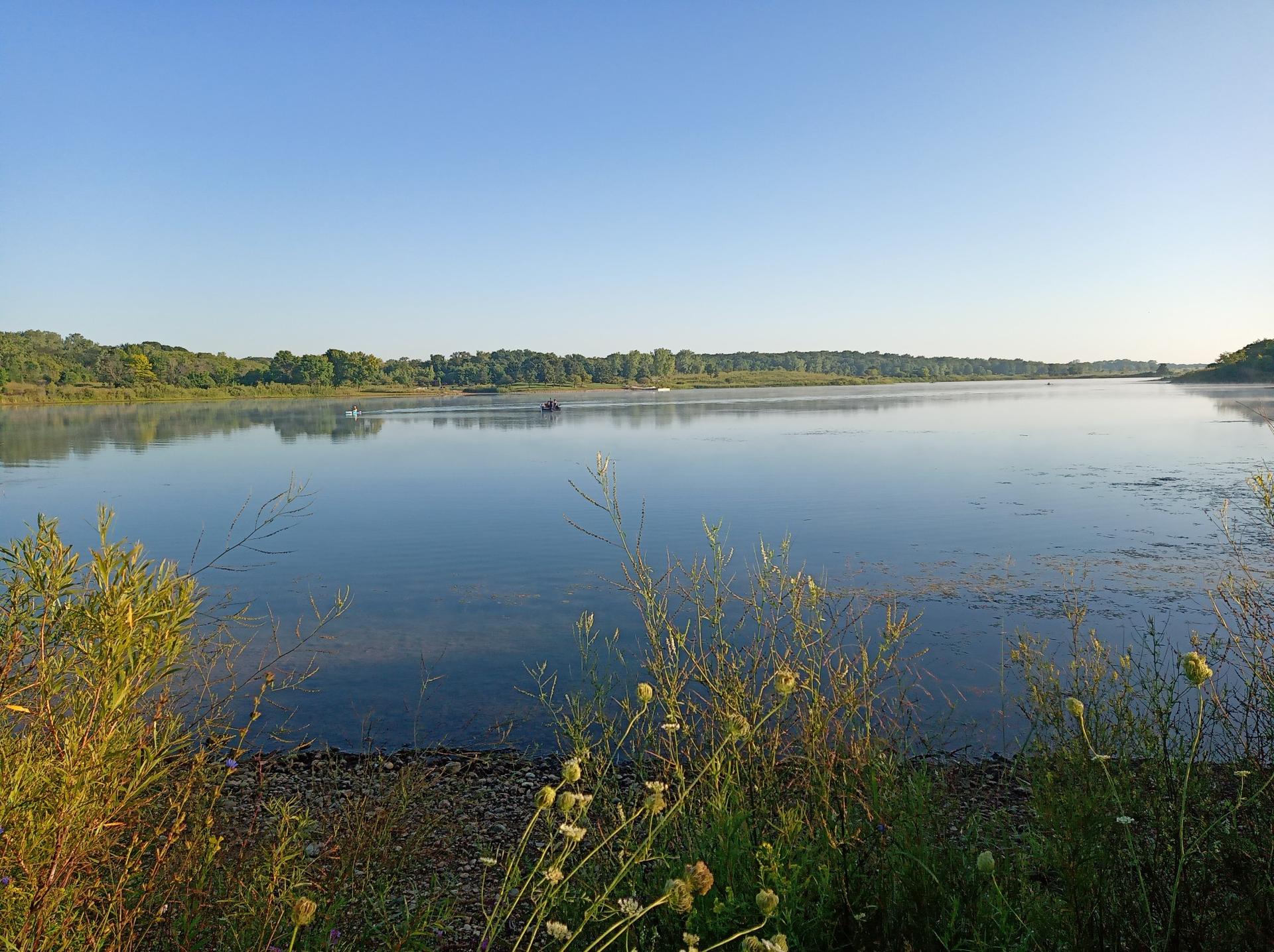 Wide calm lake under clear blue sky, distant treeline, sunny morning.