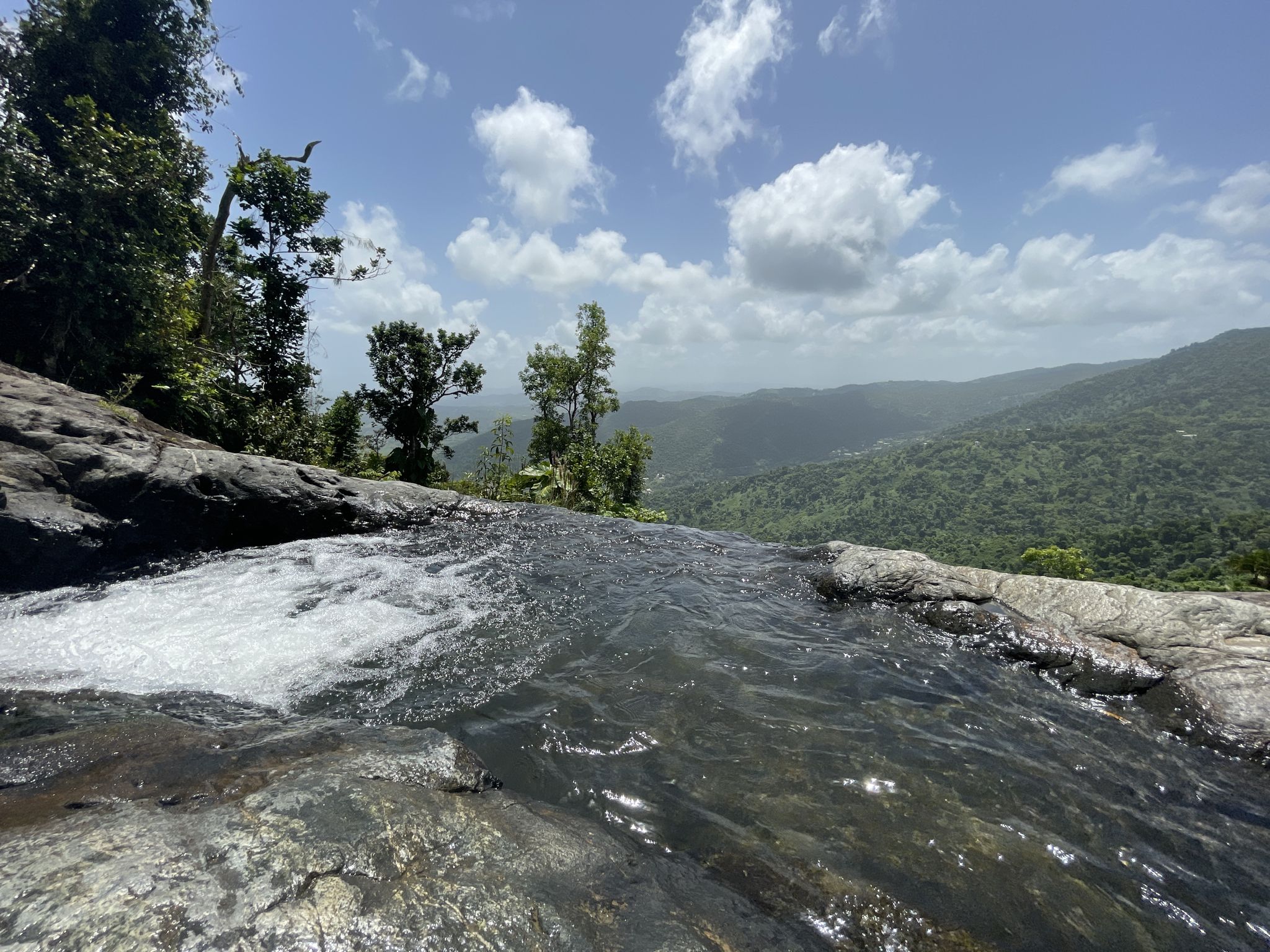 Water cascades over rocks with lush green mountains under a bright sky. - Naguabo Infinity Pool Trail