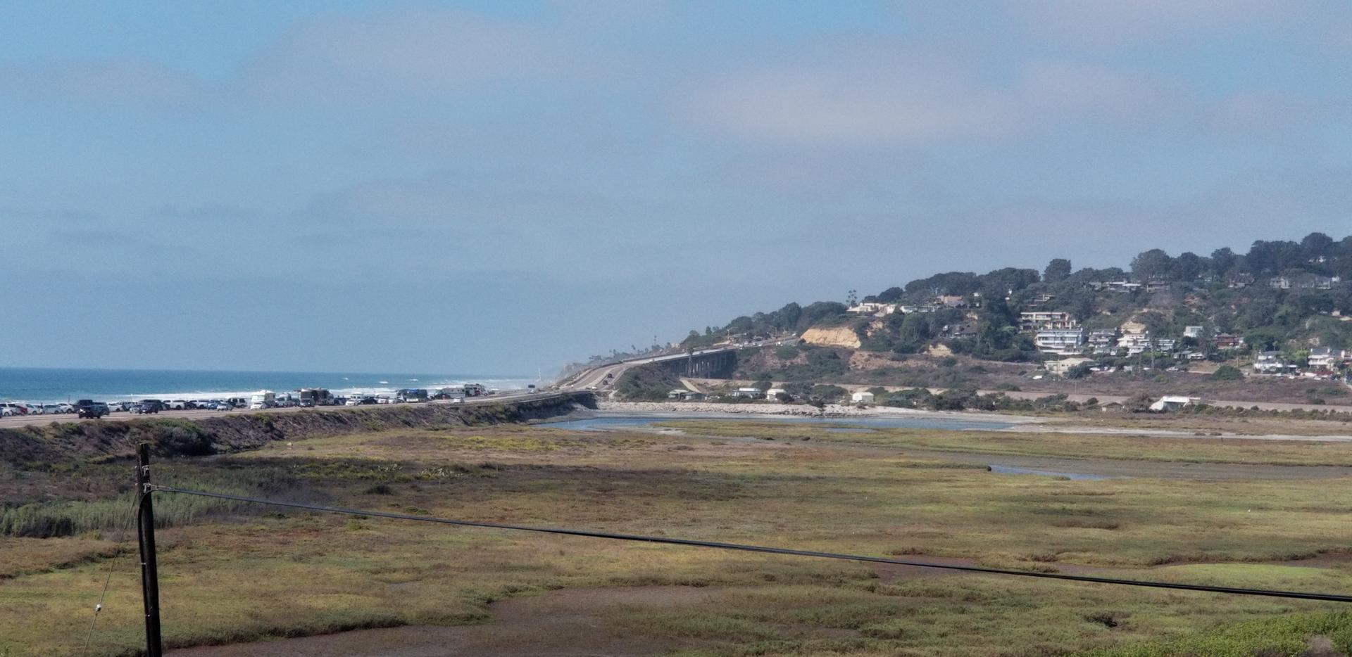 Sunny coastal view with marshland, distant ocean, highway, and hillside homes.