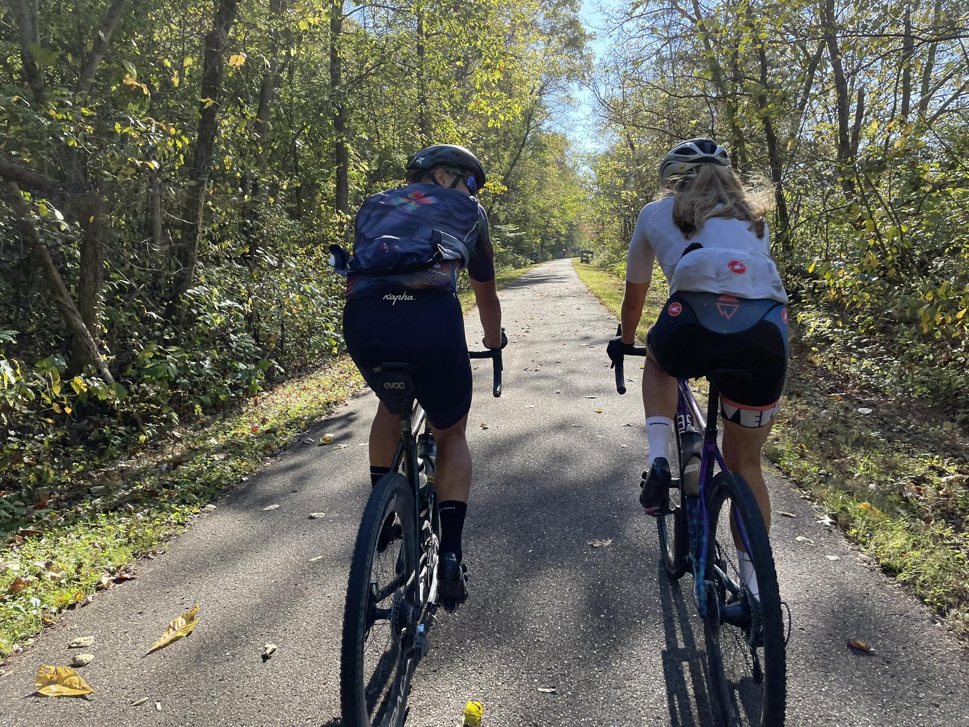 Two cyclists ride away on a sunny paved path through green trees.