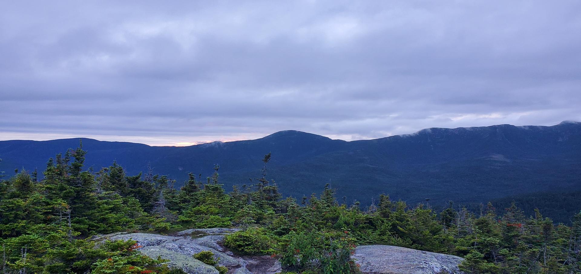 Overcast mountain range with evergreen trees and rocky foreground under a cloudy sky.