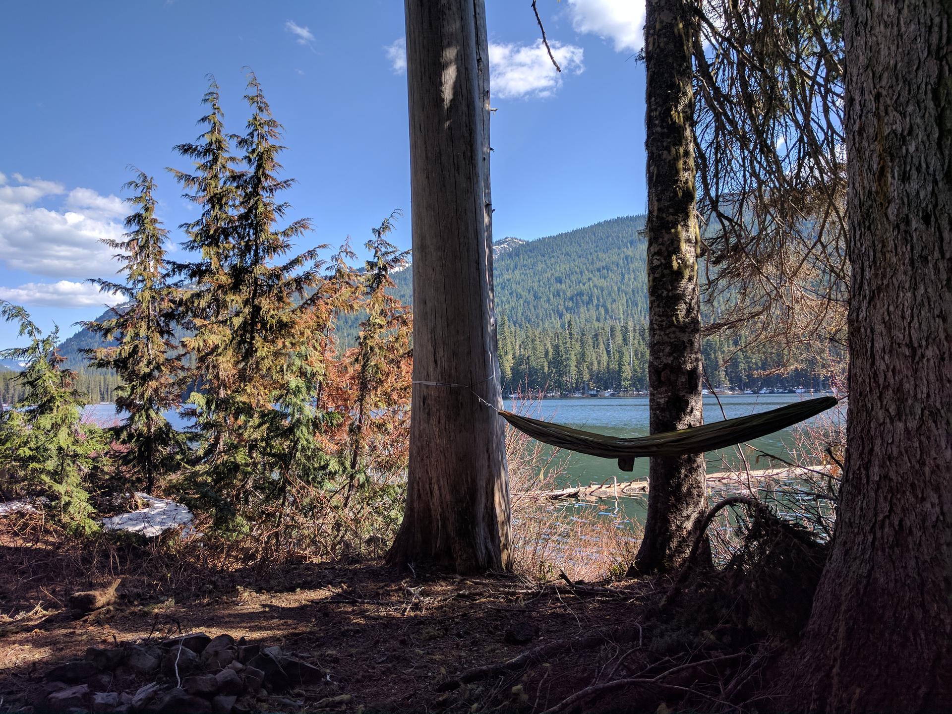 Green hammock tied between trees overlooking a sunny lake and mountains.