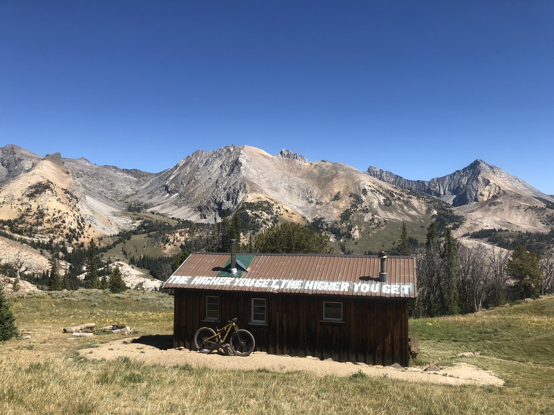 Wooden cabin with a bicycle in a dry mountain landscape under a clear sky.